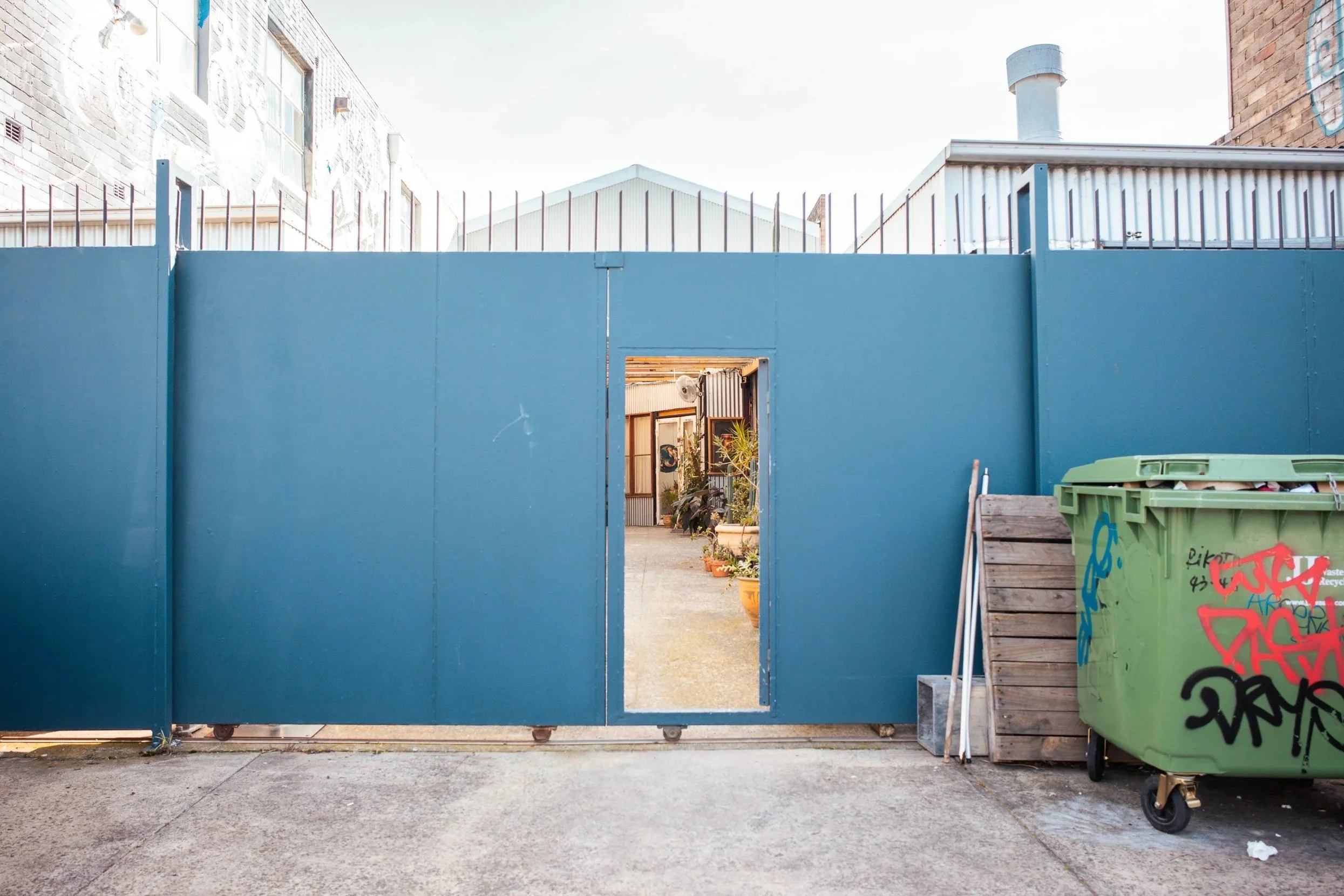 A blue metal gate with a rectangular opening in the center, revealing a courtyard with potted plants and building walls with artwork. On the right, there is a green trash bin with graffiti, leaning against wooden pallets and metal rods. The ground is