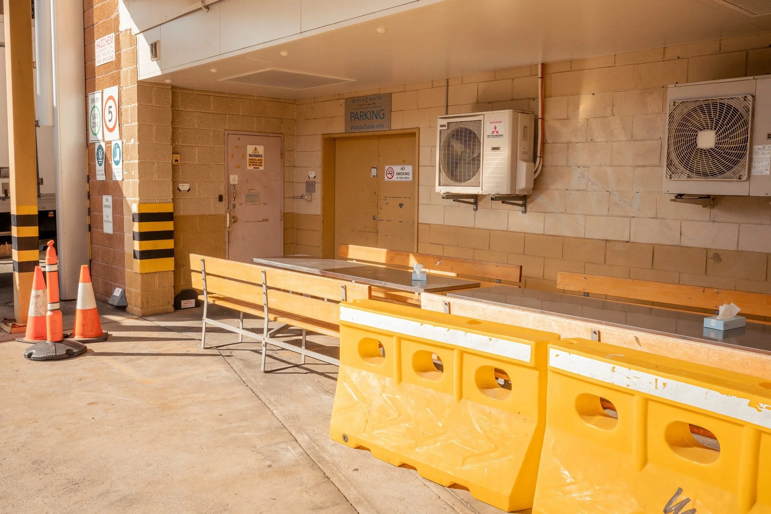 Empty outdoor seating area with a long table and benches, orange traffic barrels, and yellow safety barriers outside a building with parking and no smoking signs, air conditioning units, and a door.