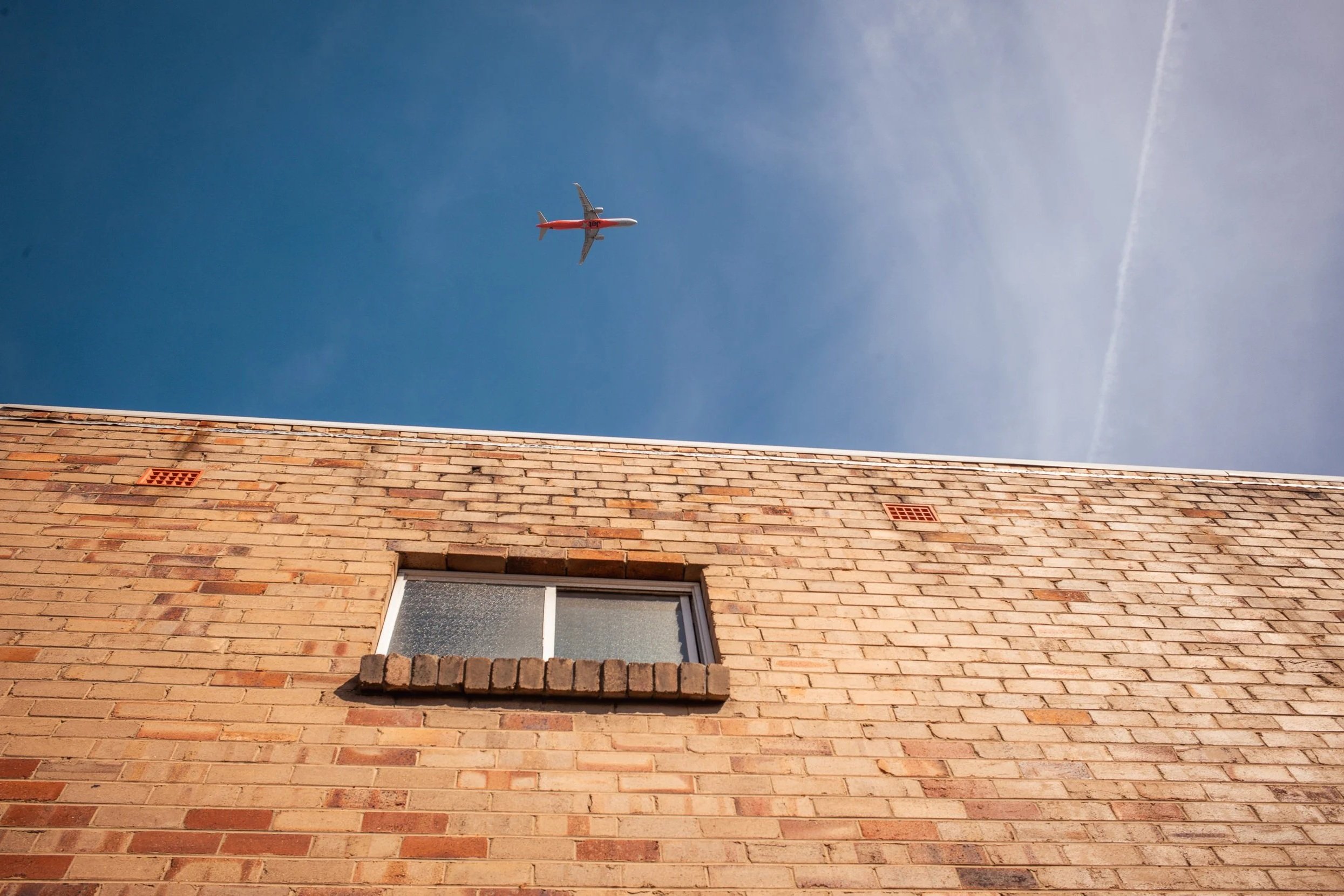 View of a clear blue sky with an airplane flying above a brick building with a window.
