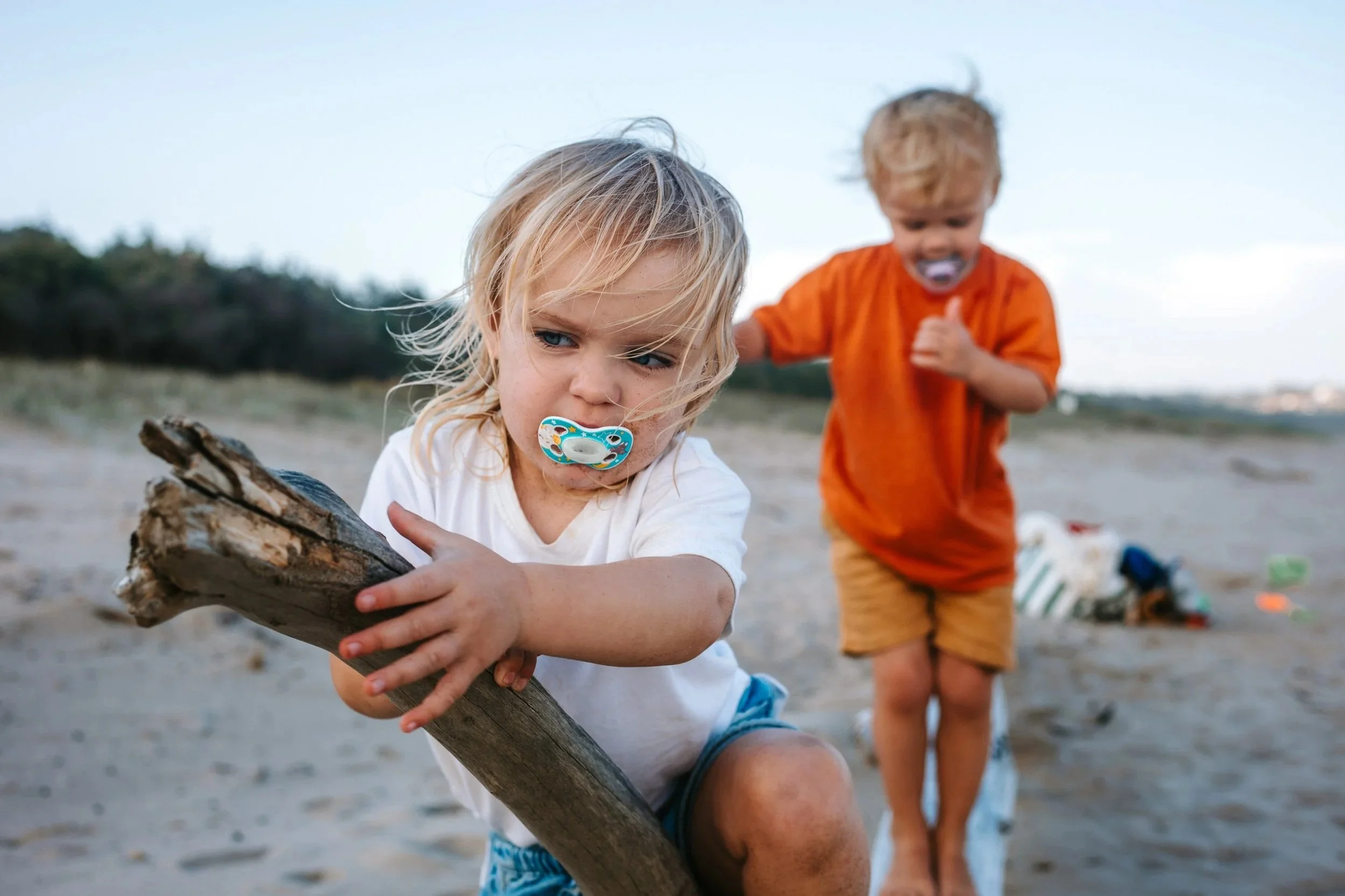 Two young children playing on the beach, one girl with a pacifier holding a large piece of driftwood, and a boy in an orange shirt and beige shorts walking nearby, with beach toys and sand in the background.