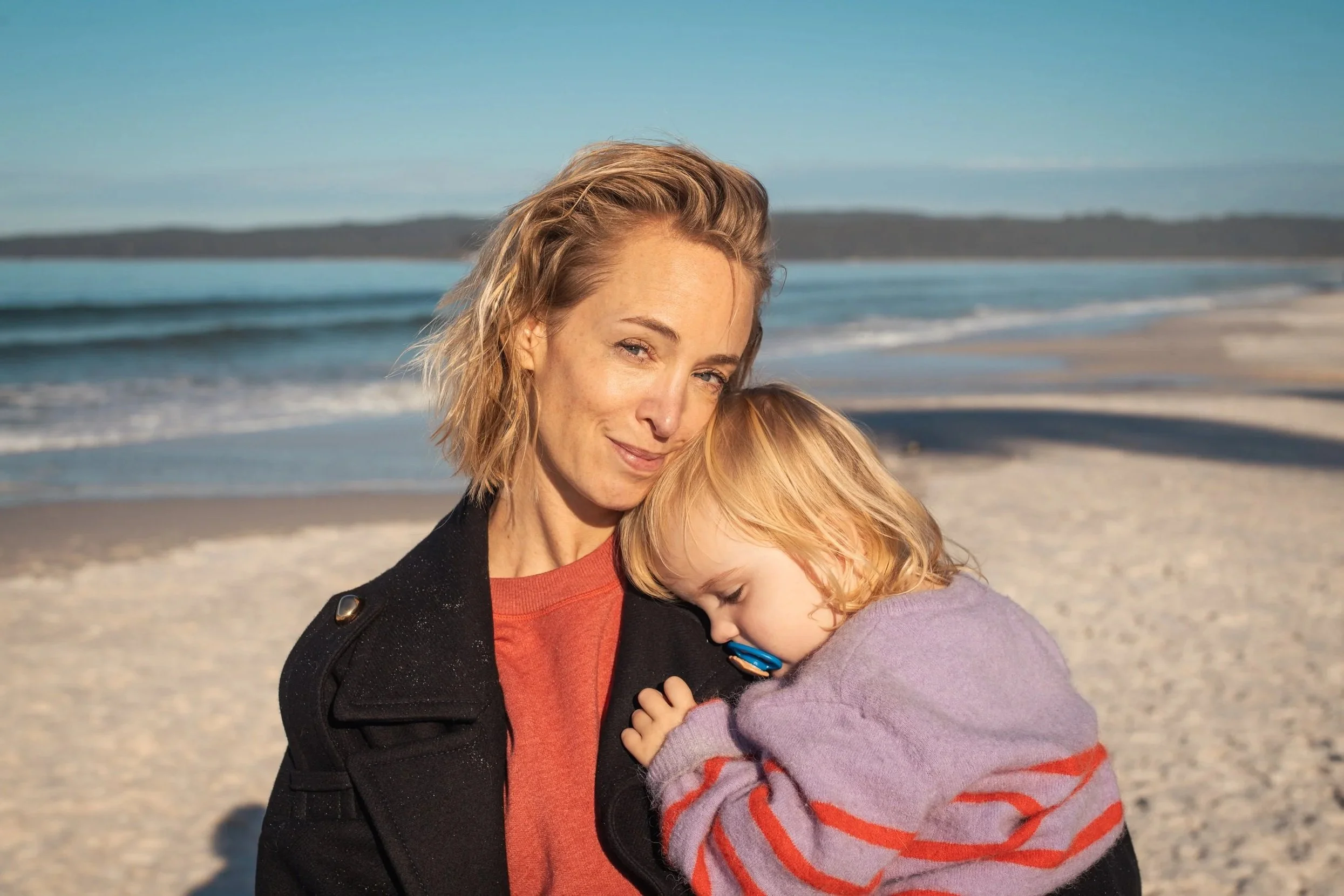 A woman and a young girl hugging on a beach with ocean waves and a distant shoreline in the background.