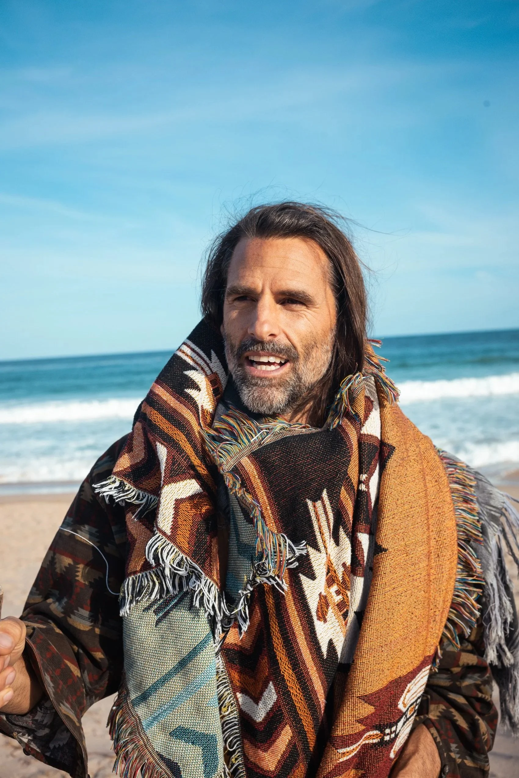 A man with long hair and a beard at the beach, wearing a patterned blanket or shawl.