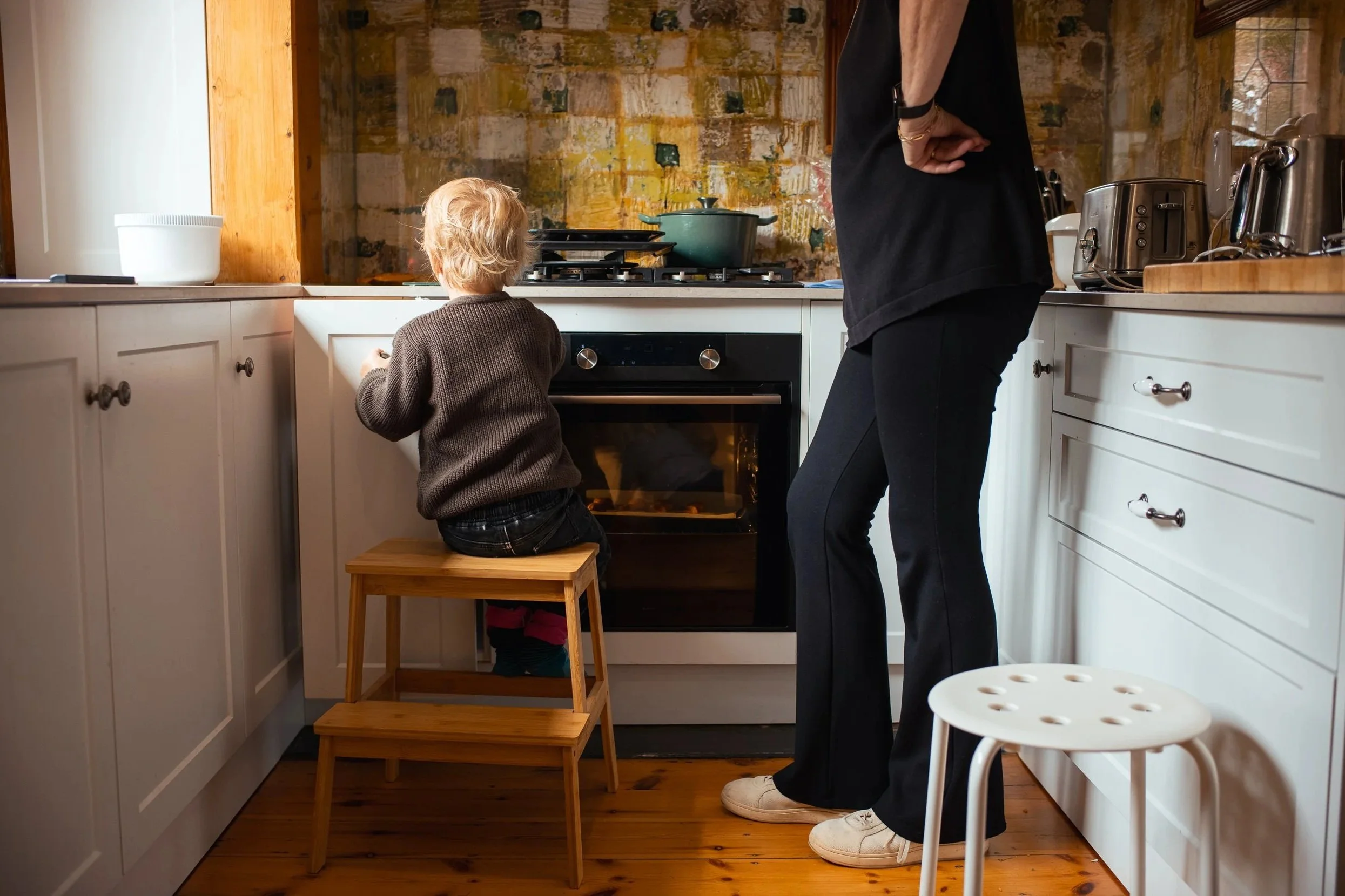 A young child sitting on a wooden stool and looking into an oven in a cozy kitchen, with an adult standing nearby.