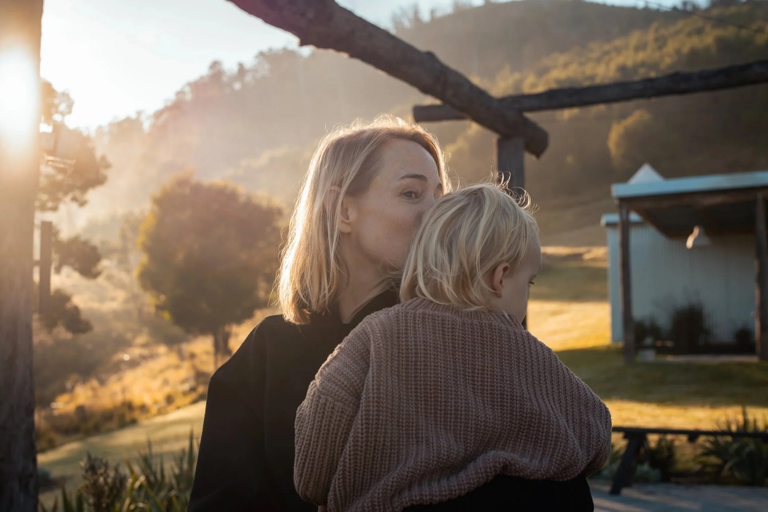 A woman holding a young child outside during sunset or sunrise in a rural or park setting with trees and rolling hills in the background.