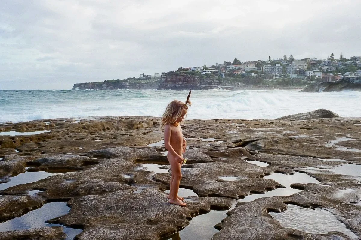 A young girl with blonde hair standing on a rocky beach near the water, looking down with her hand in front of her, with houses on a cliff in the background under a cloudy sky.