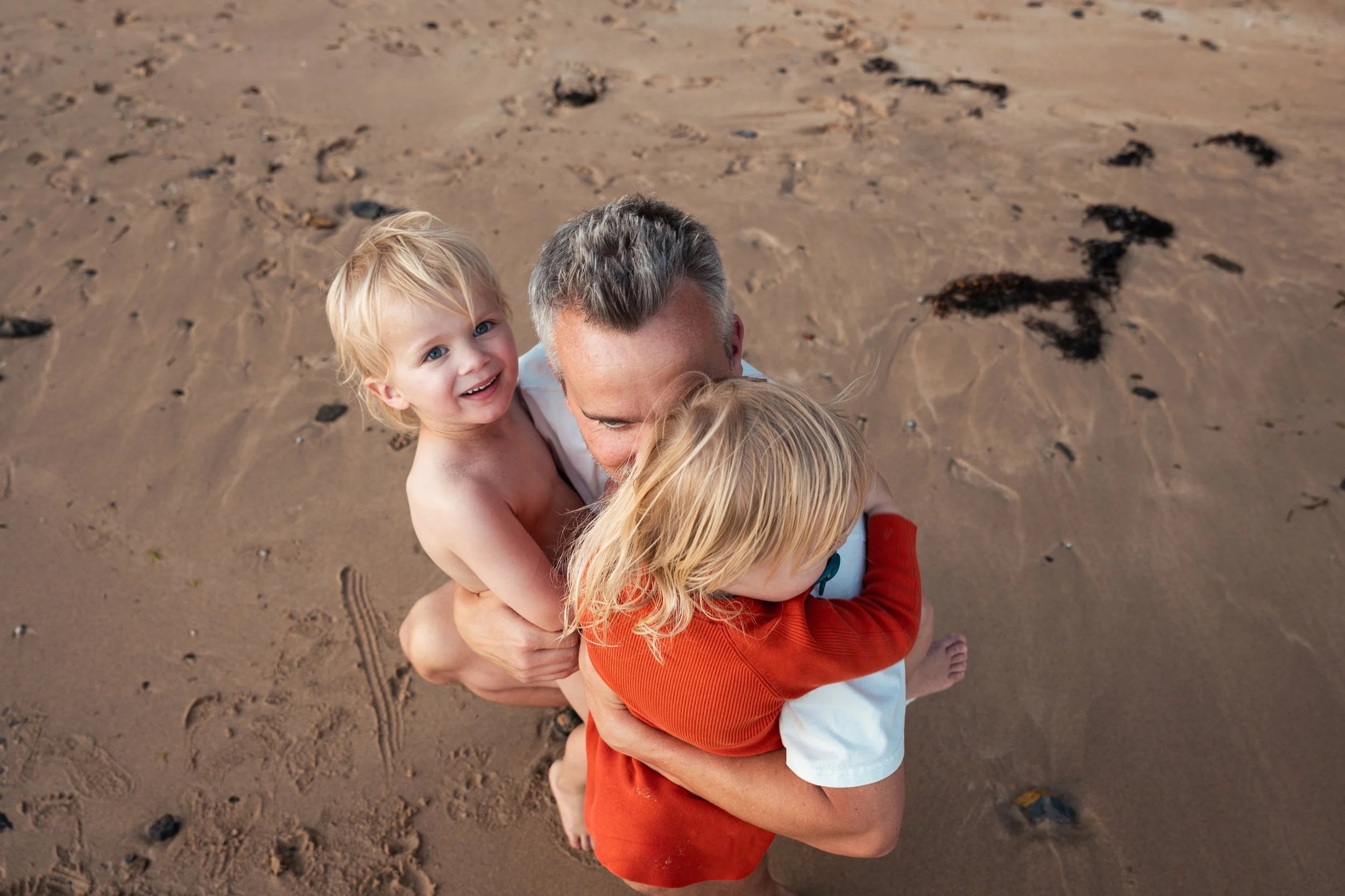 A man hugging two young children on a sandy beach, one child smiling and the other with their head down.