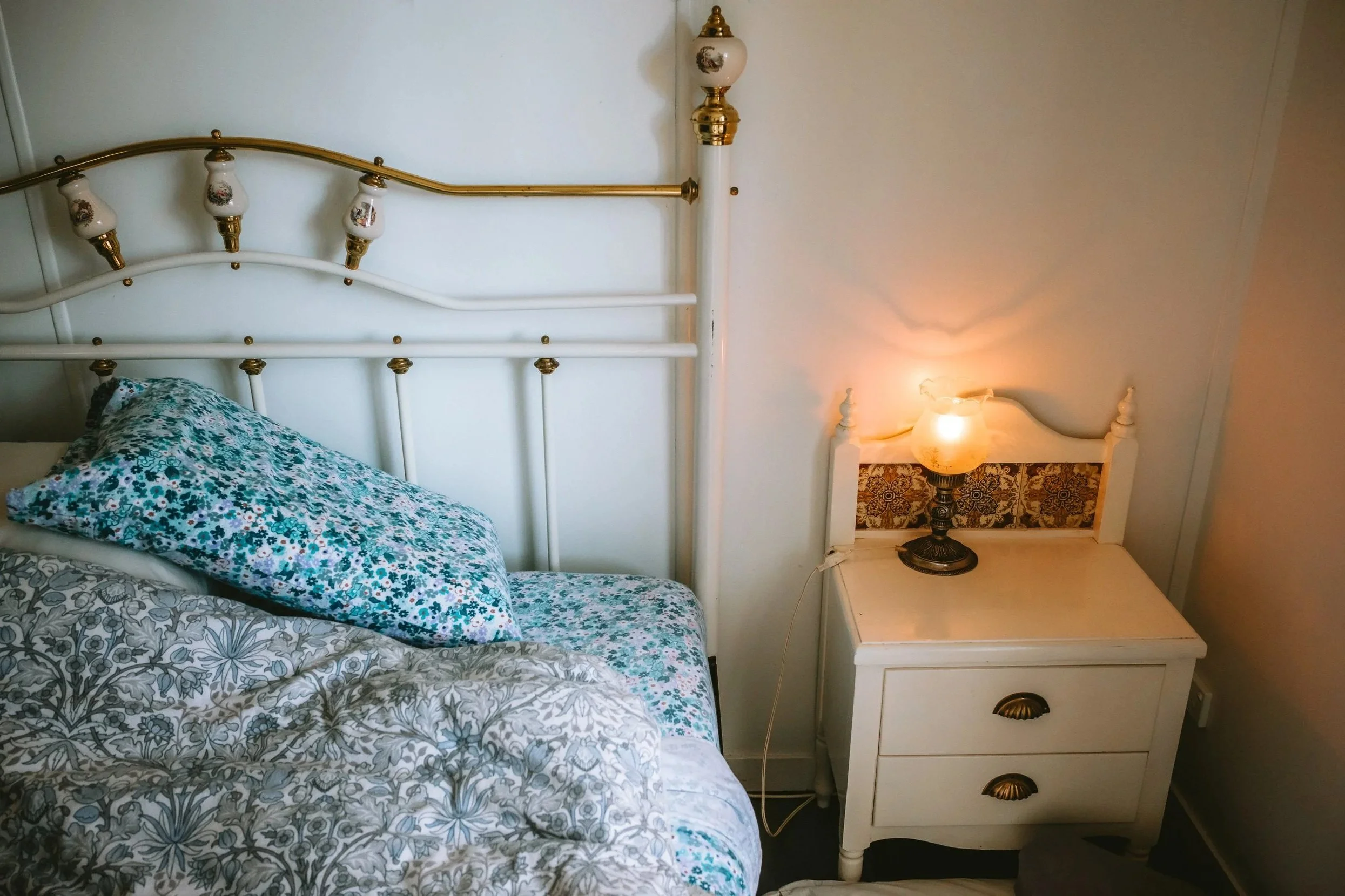 A bedroom with a white metal bed frame with gold accents, a floral pillow, and a white nightstand with a vintage-style lamp.