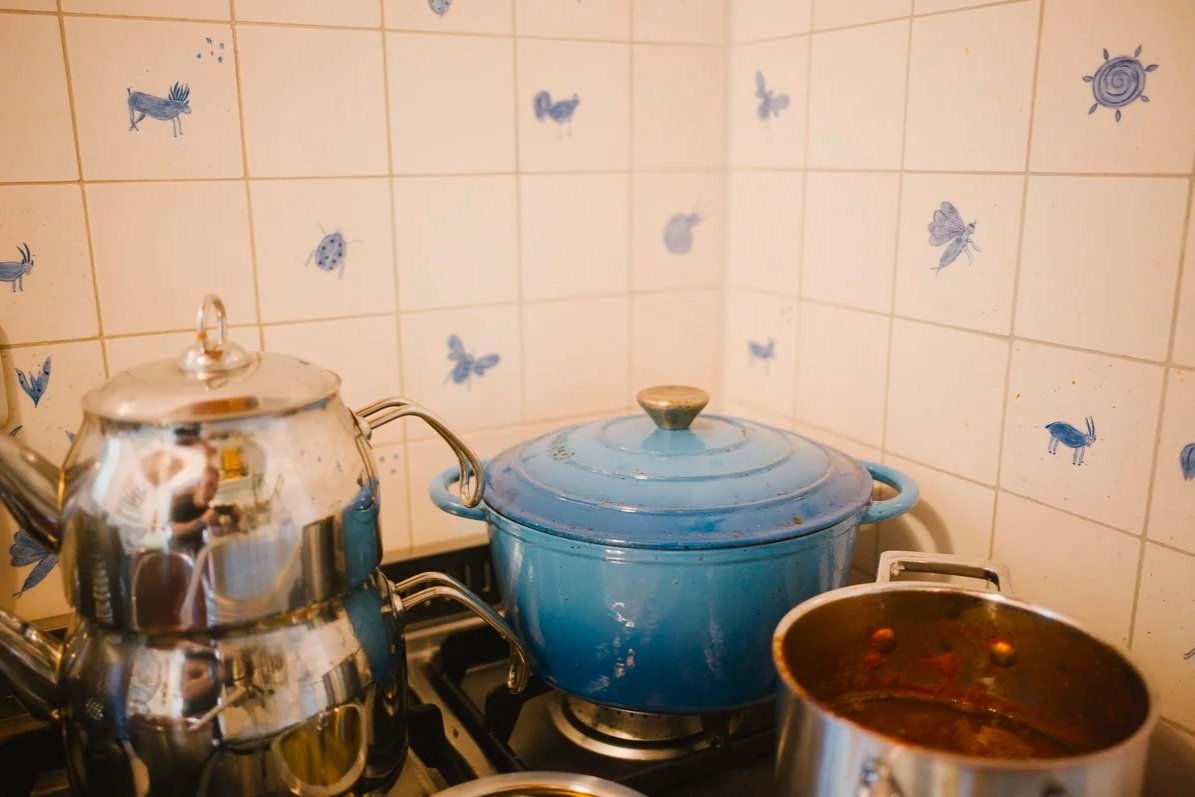 Kitchen stove with a silver kettle, a blue pot with a lid, and a silver pot with a reddish sauce. The background has tiled walls with blue bug and insect stickers.