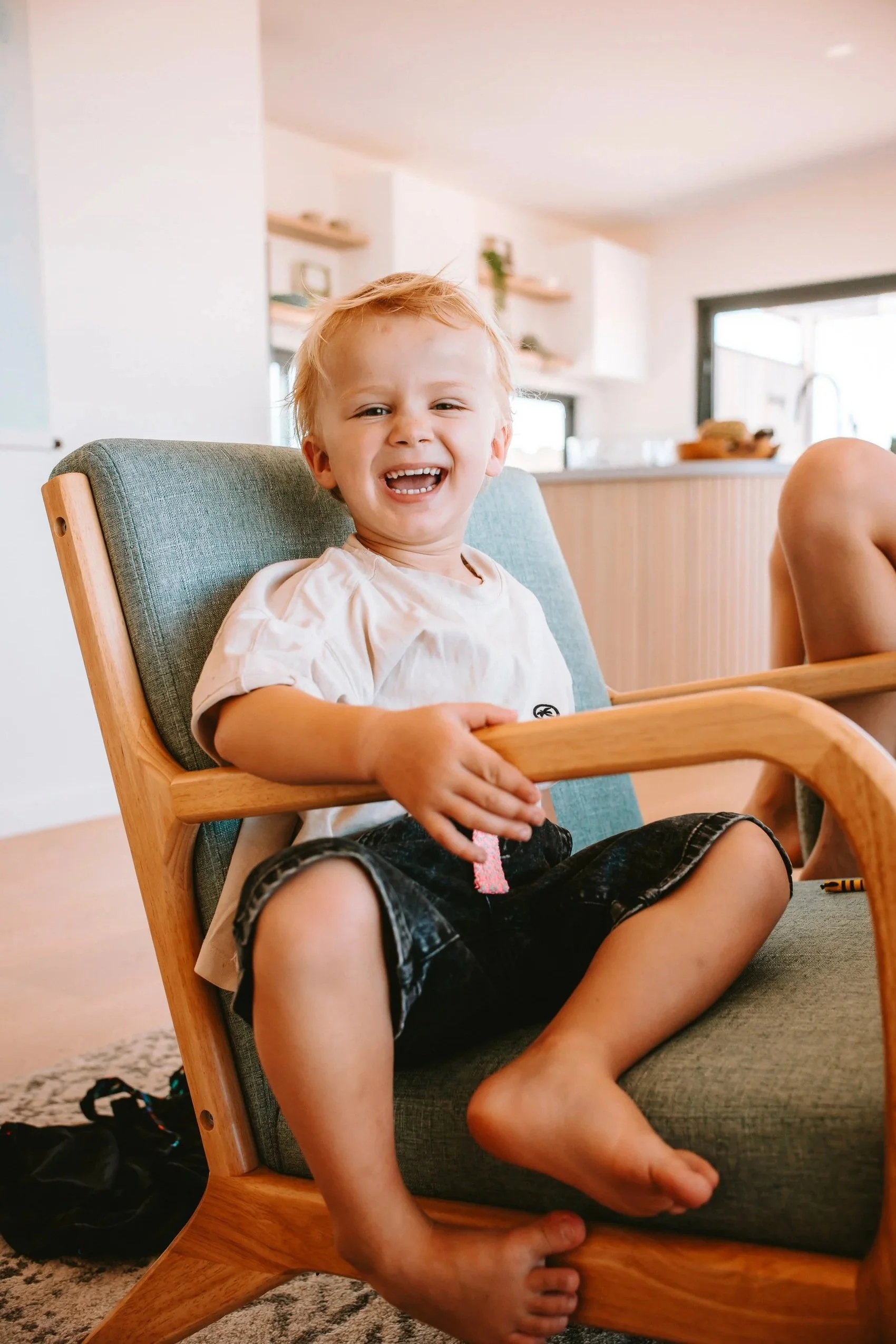 A young boy sitting in a modern gray and wood armchair, laughing and smiling widely in a bright, modern kitchen and living space.