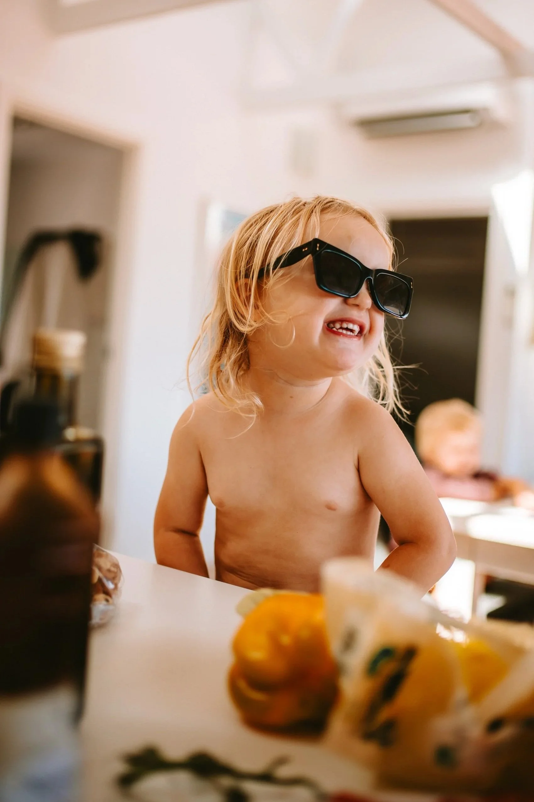 A young child with long blonde hair, wearing sunglasses, and no shirt, smiling in a kitchen with autumn-themed decorations.