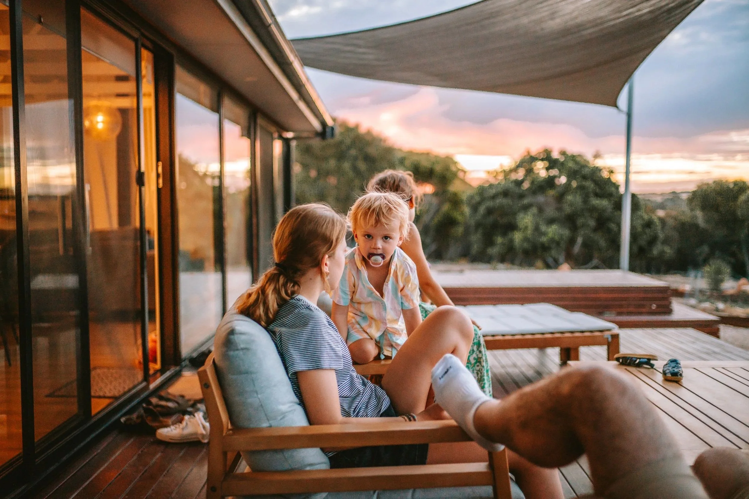 Parents and children sitting on a wooden deck during sunset, with trees and a colorful sky in the background.