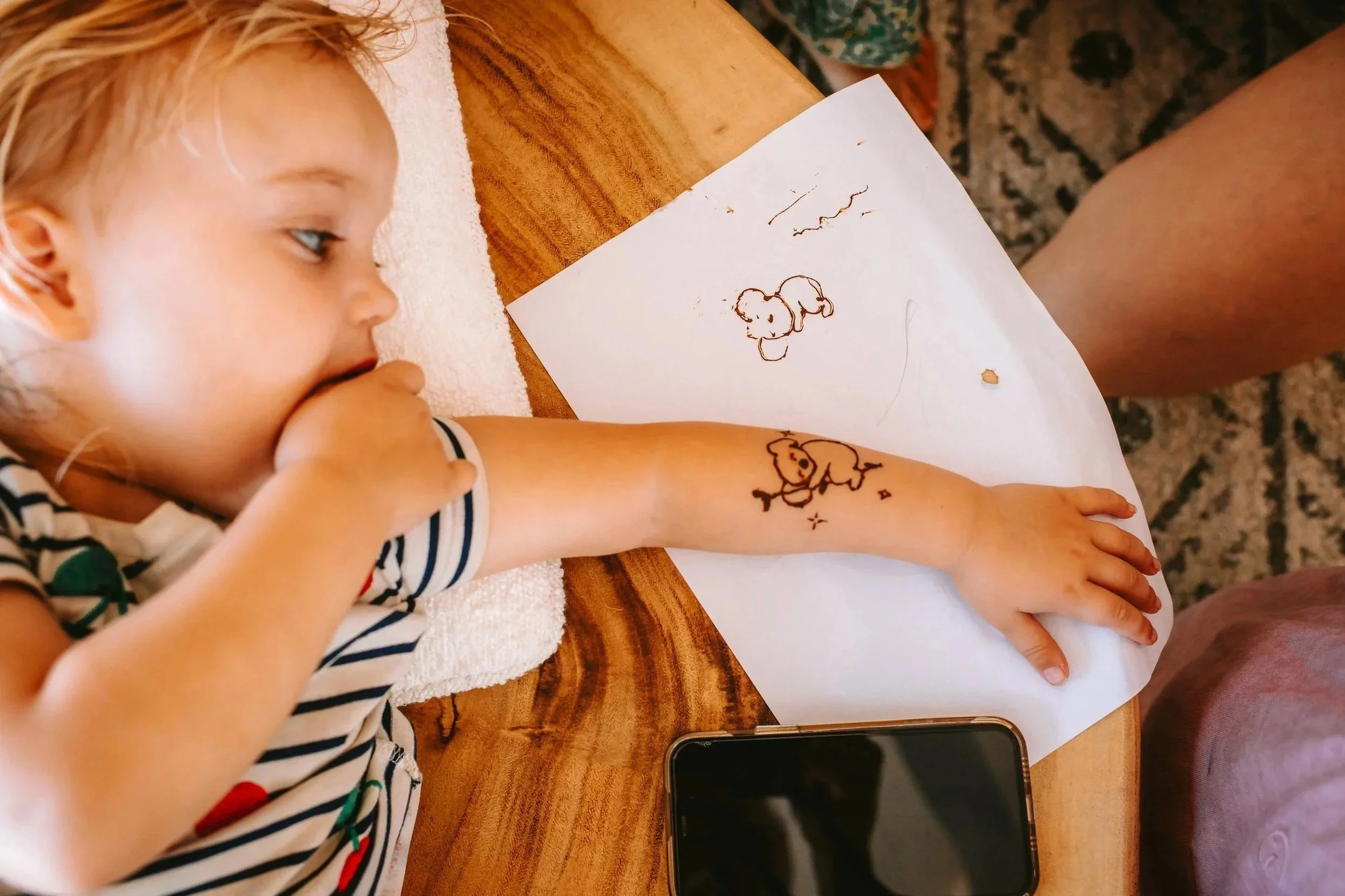 A young child lying on a table with a towel beneath their head, resting their head on one hand. The child's arm is extended, showing a temporary tattoo of a dog holding a stick. There is a piece of paper with dog drawings and some scattered marks on 