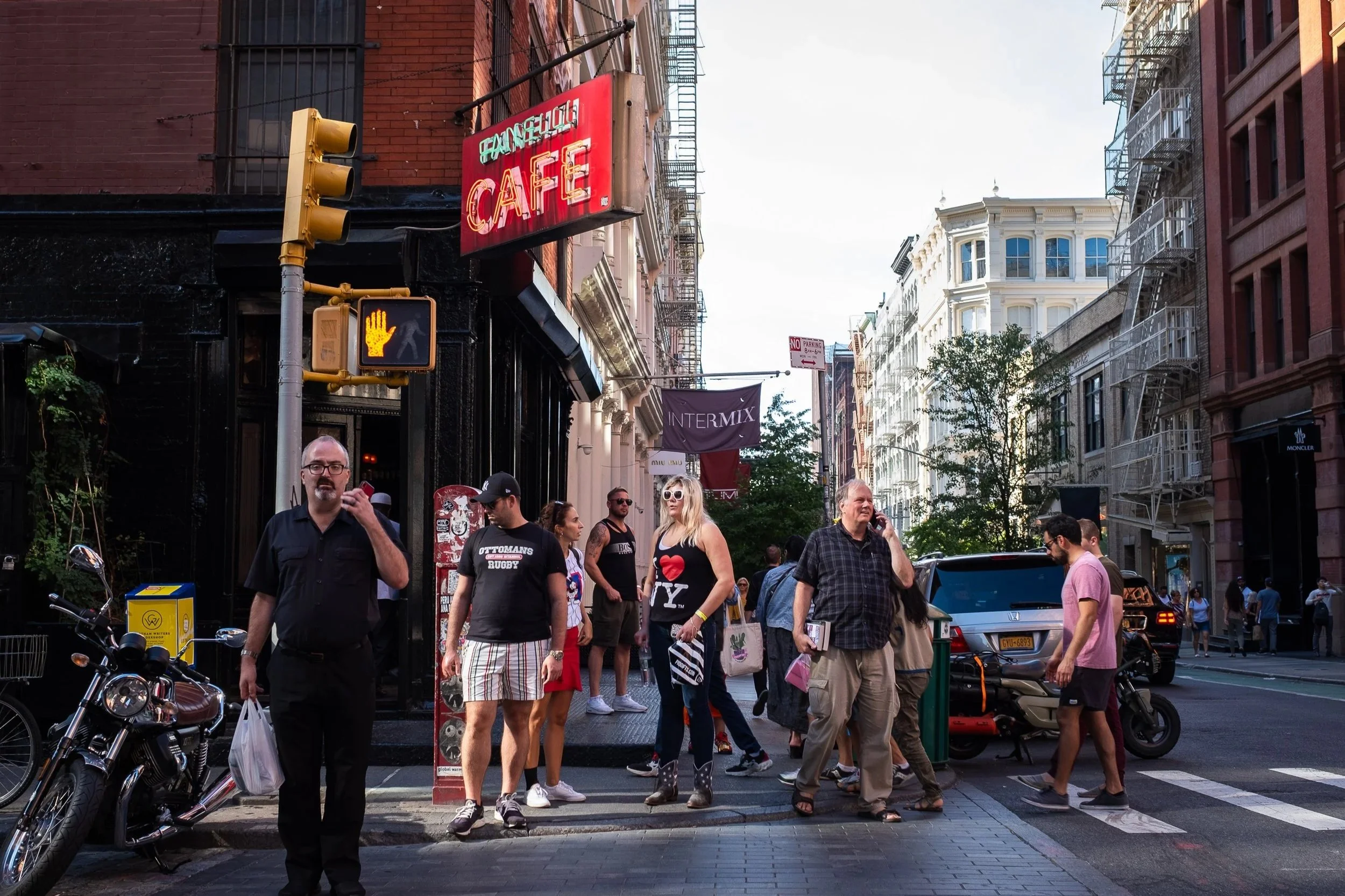 City street scene with several pedestrians waiting at a crosswalk, including a man in black, a woman in sunglasses, a man with a checkered shirt, and others. There is a motorcycle parked on the sidewalk, a traffic light showing a yellow hand signal, 