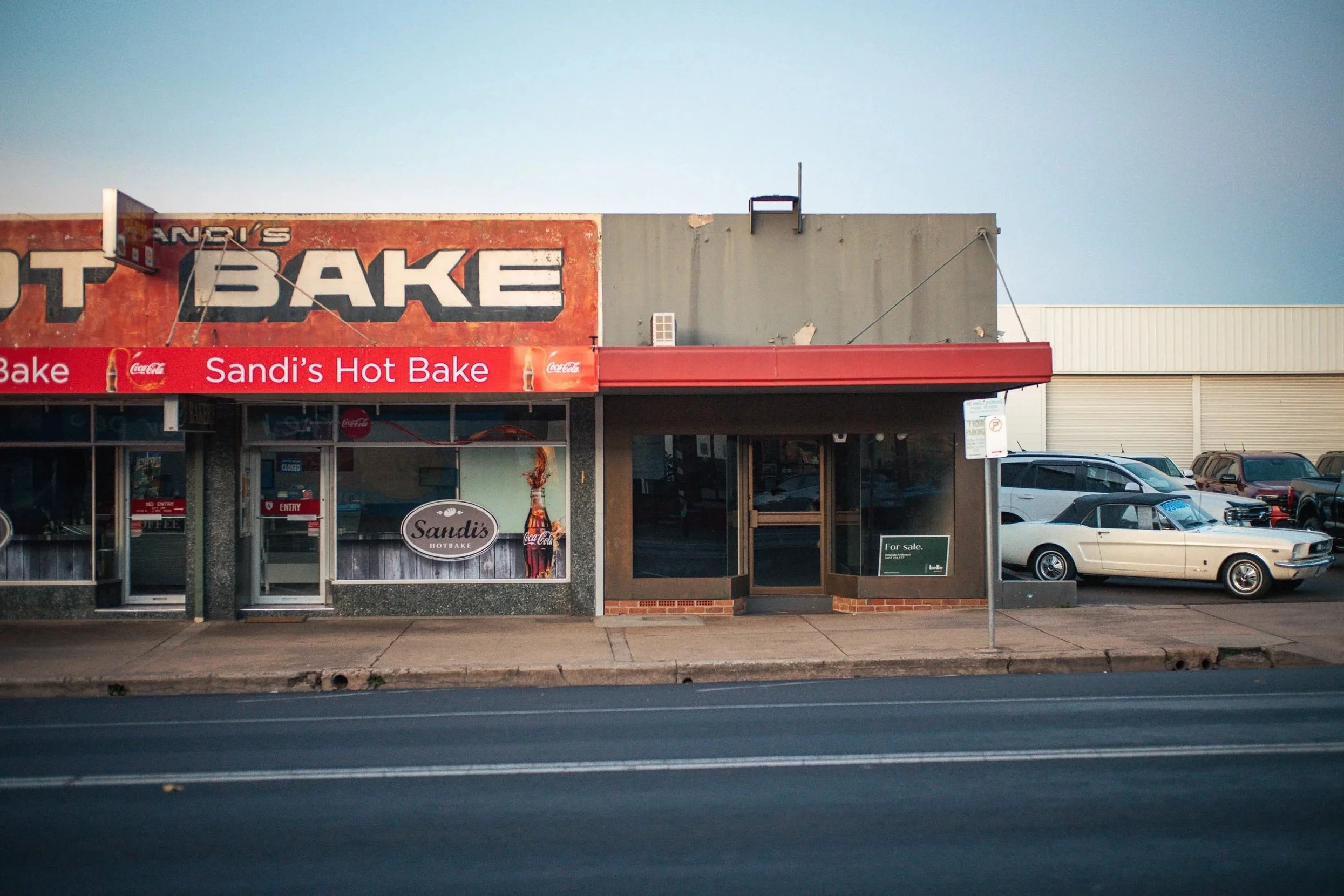 Front view of a small storefront with a red sign reading "Sandi's Hot Bake" and a Coca-Cola advertisement with a soda bottle and glass. The building has glass doors, one labeled "Entry," and large windows. There are parked cars on the street and a si