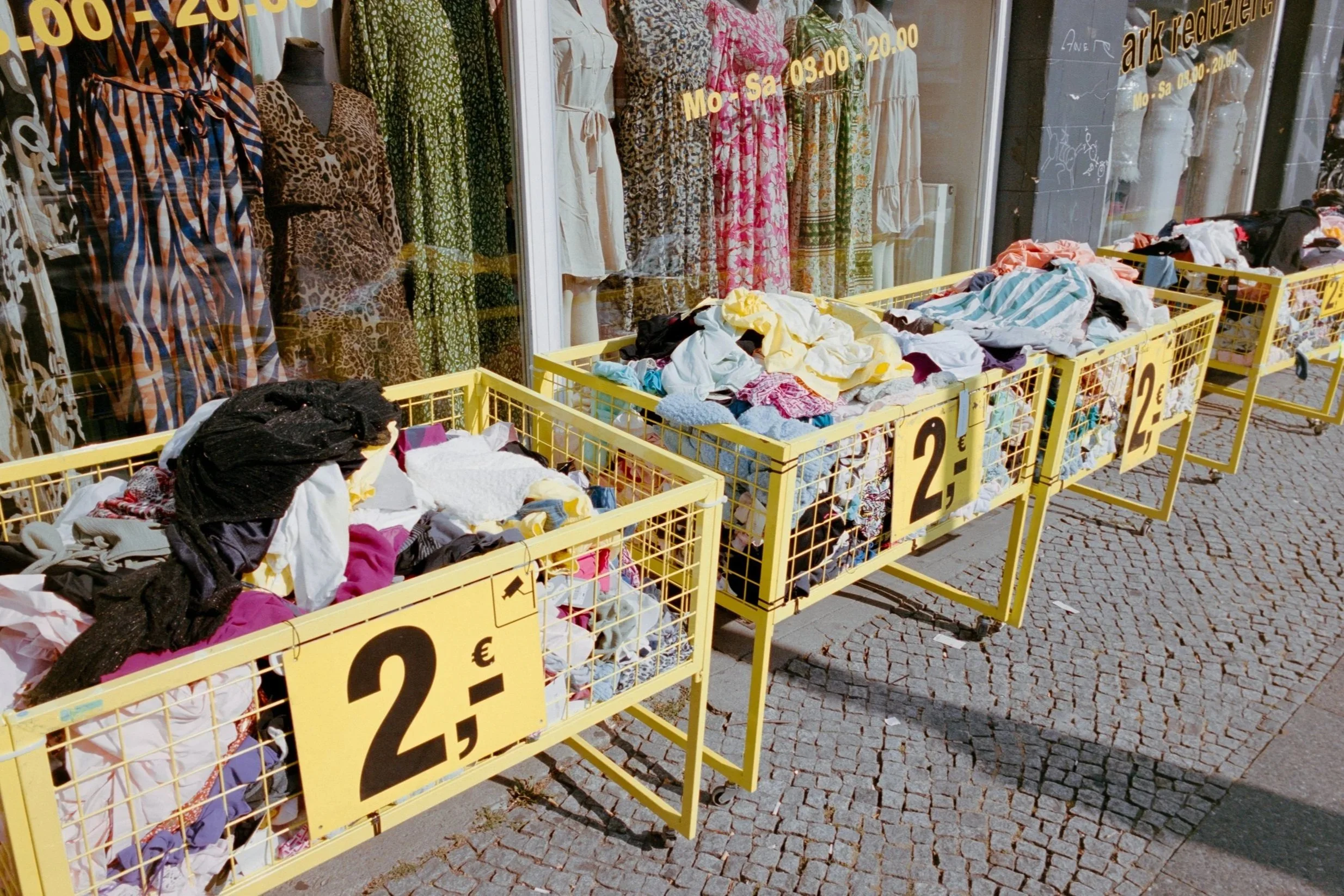 Outdoor scene with yellow shopping carts filled with clothes, lined up on a cobblestone sidewalk outside a store with mannequins and dresses in the window. Signs on carts indicate a price of €2.50.