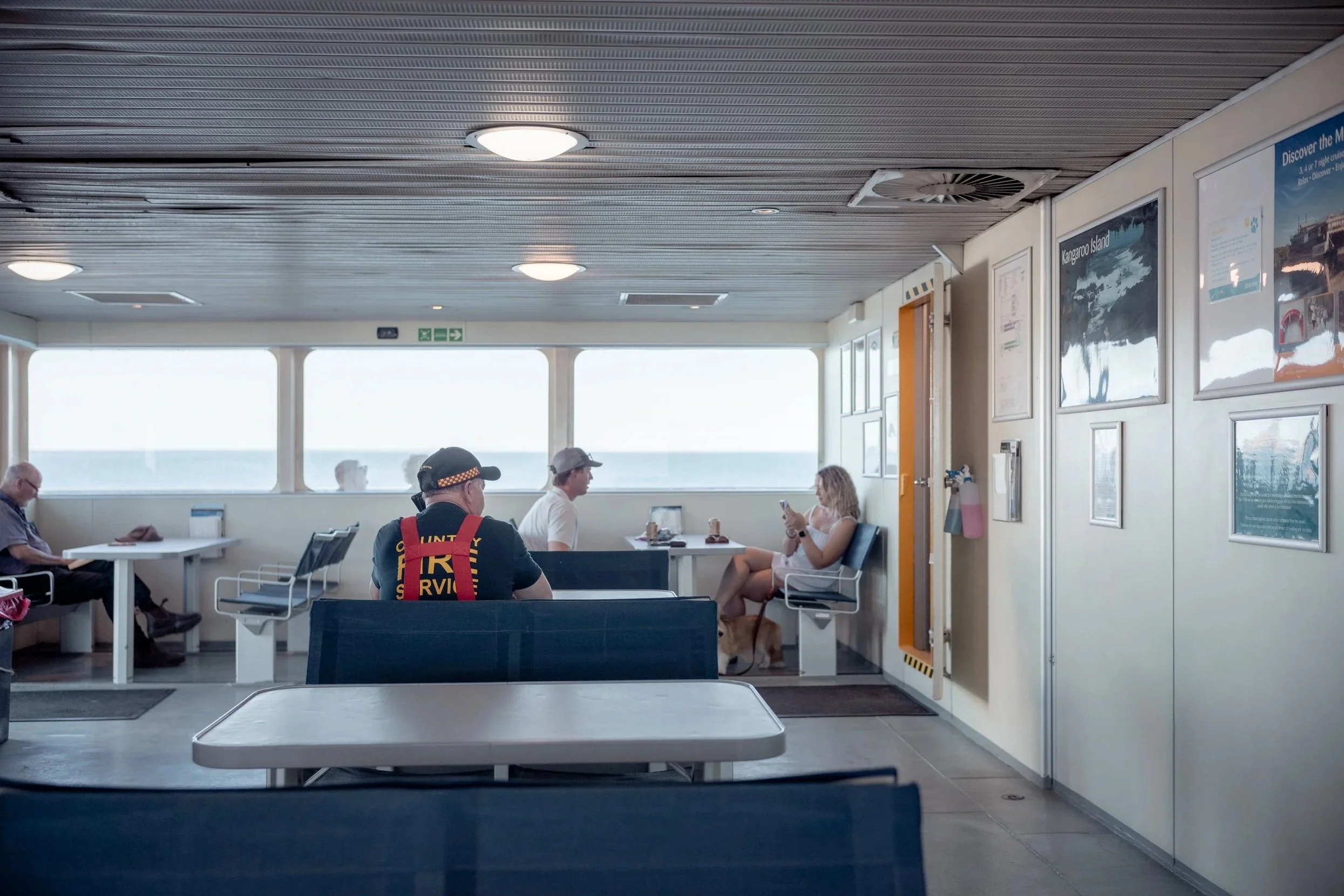 People sitting in an indoor ferry or cruise ship lounge with large windows showing the ocean outside. Some are using their phones, and one person appears to be a crew member wearing a uniform with the words 'FIRE' on the back. There are overhead ligh