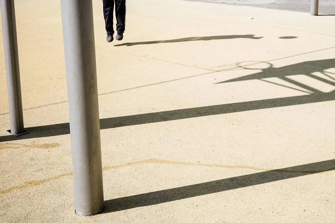 Partial view of a person's legs and feet, hanging in the air, with shadows cast on a sandy-colored ground, near metal poles.