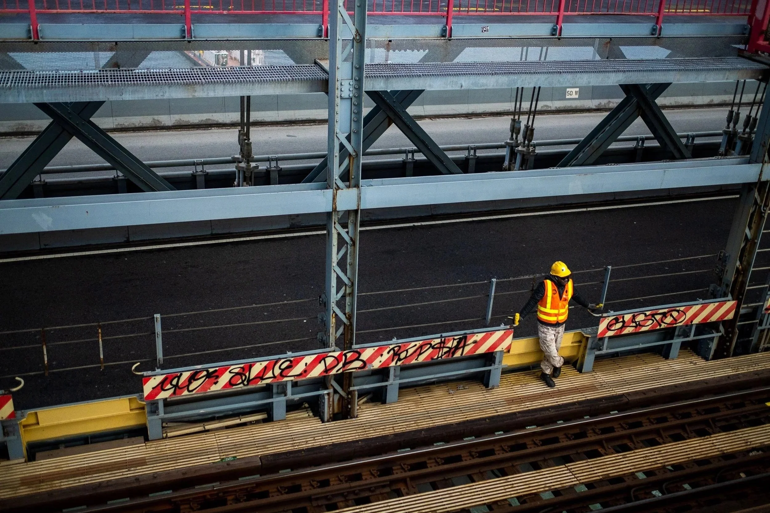 A worker in safety gear, including a yellow hard hat and reflective vest, standing on a construction or maintenance platform beside railway tracks, with construction barriers and steel structures around.