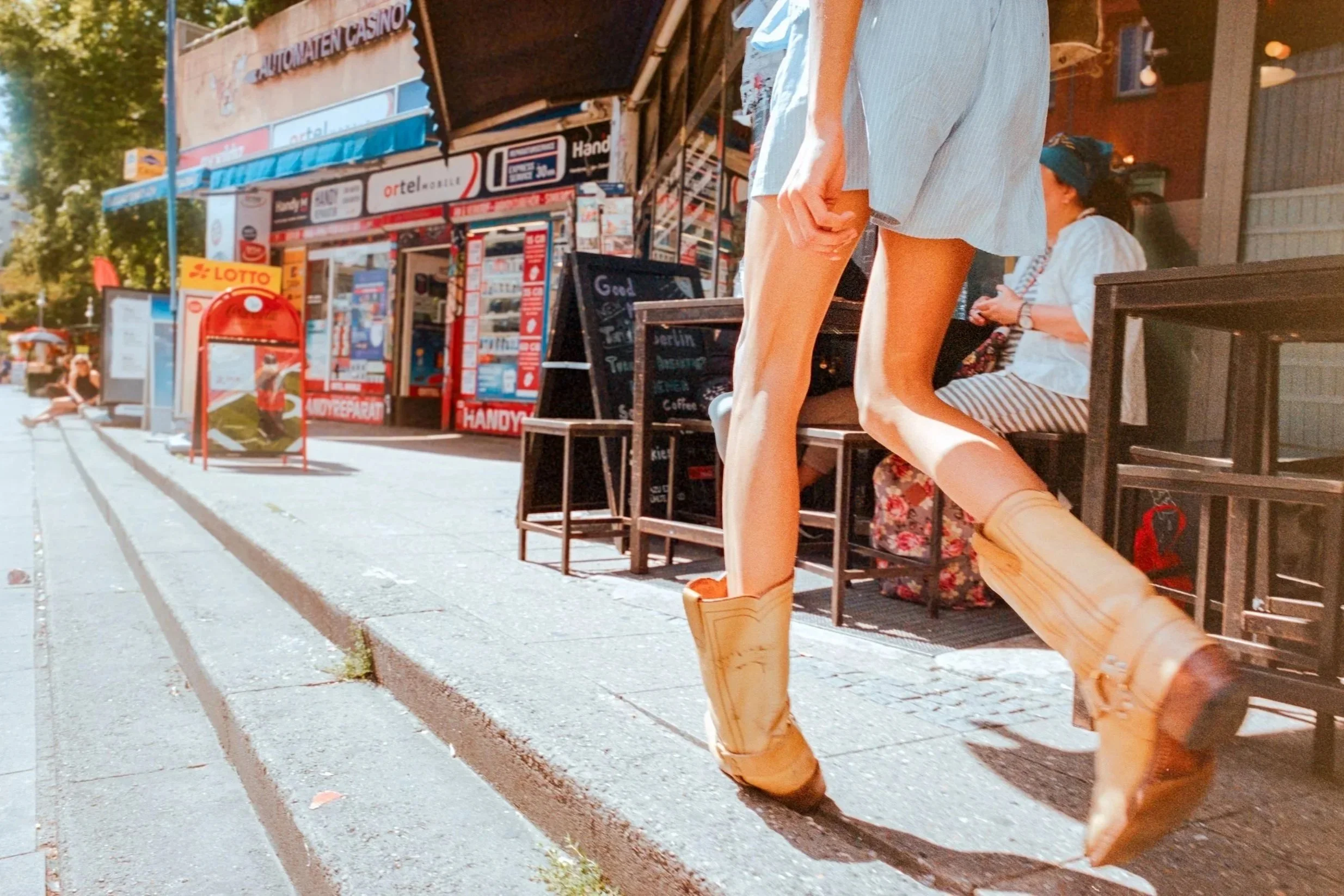 Woman walking on the sidewalk wearing tall tan boots and a light blue skirt, with a storefront in the background.
