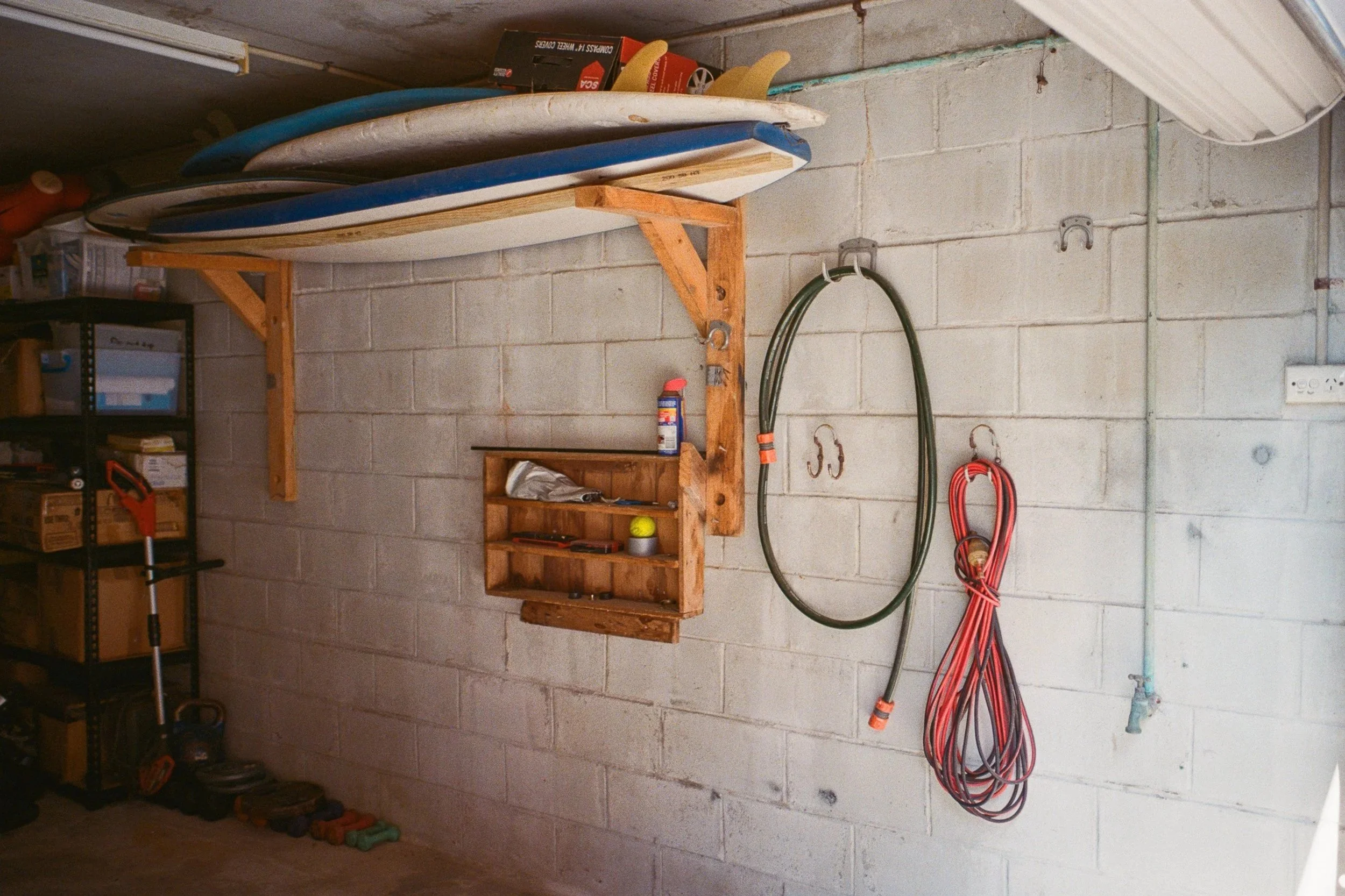 Garage with surfboards, garden hose, garden tools, and storage shelves against concrete block wall.