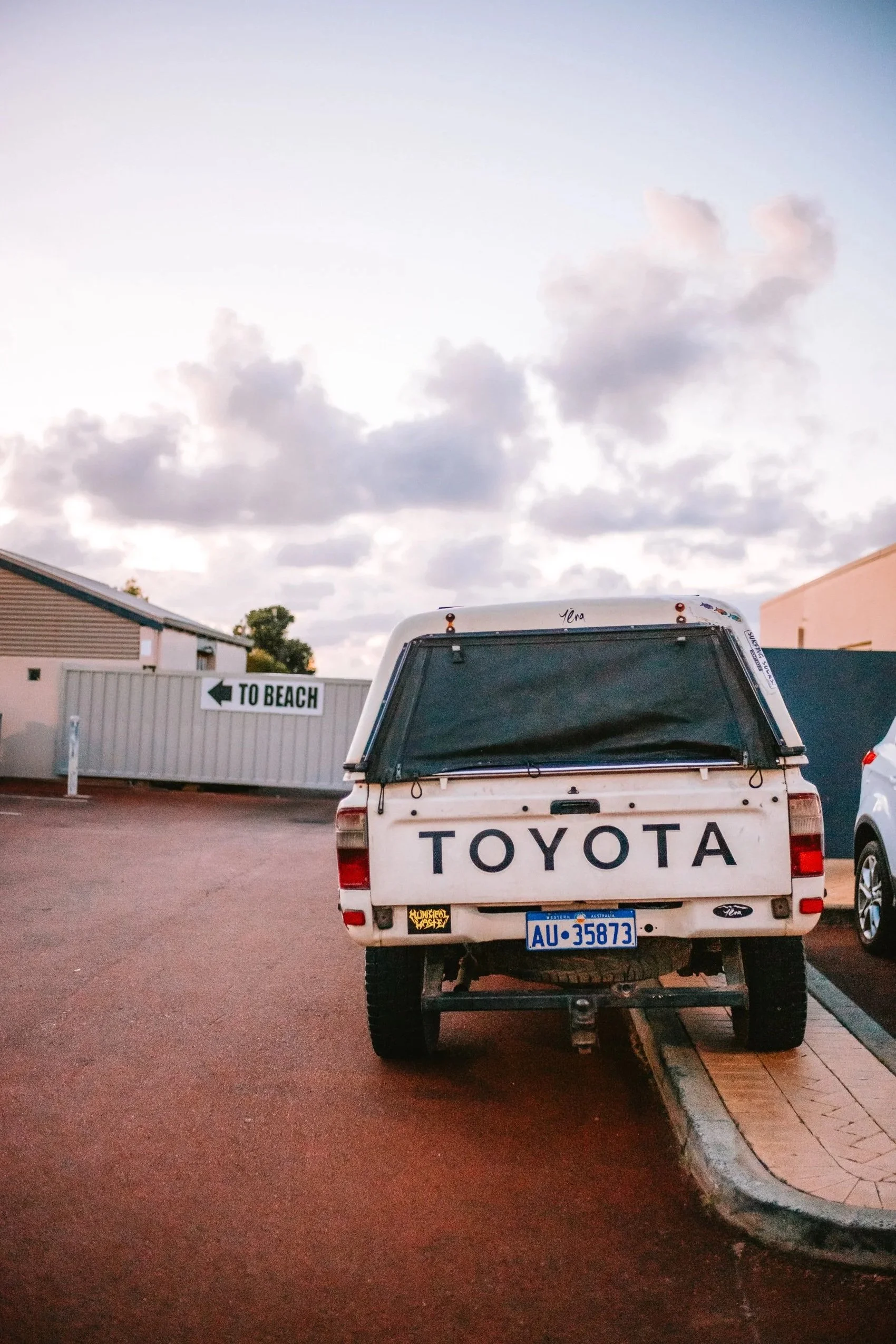 Rear view of a white Toyota pickup truck parked on a red dirt surface near a curb, with a 'TO BEAC' sign and a cloudy sky in the background.