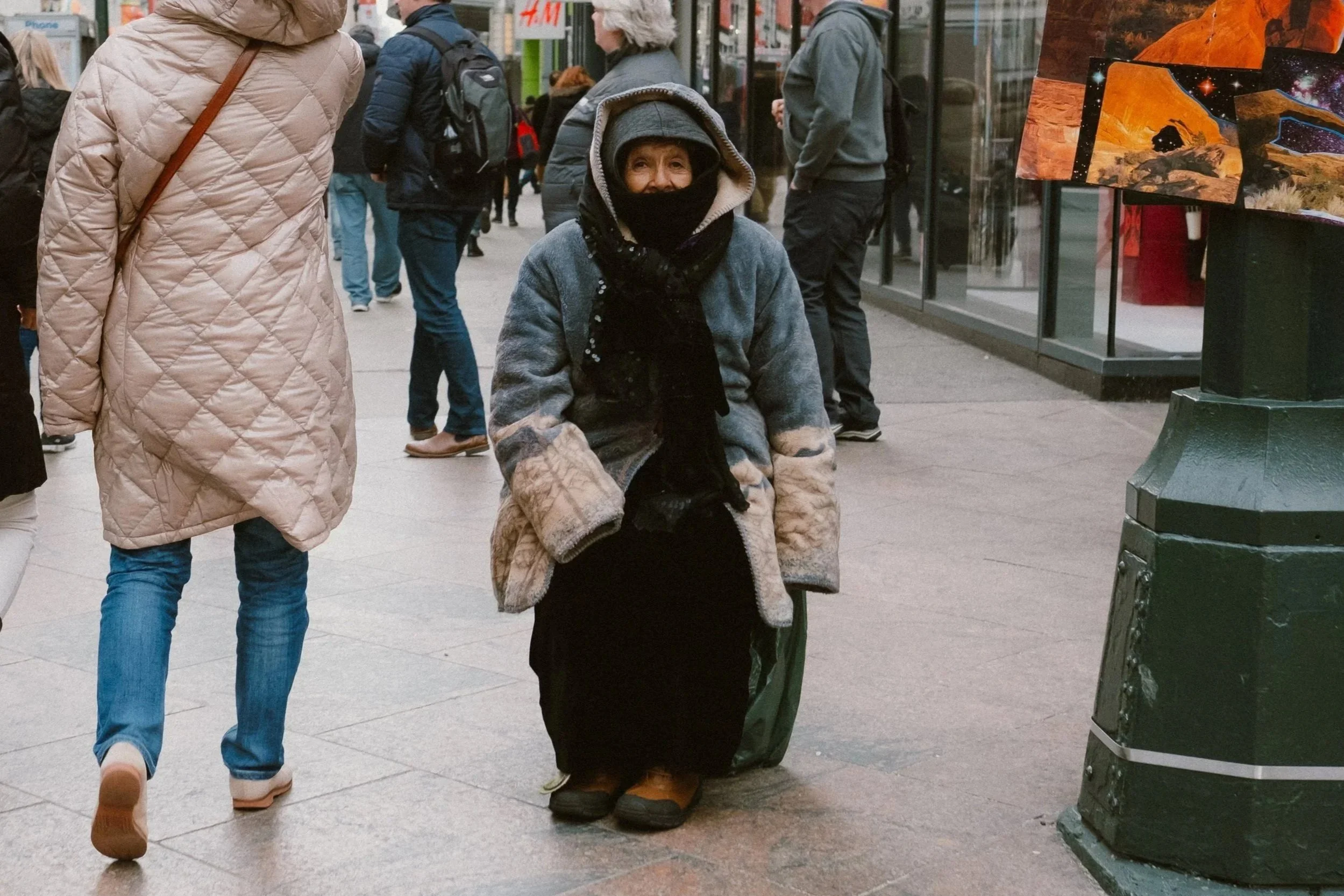 A woman sitting on the sidewalk at a busy city street, dressed warmly with a coat, head covering, and face mask, surrounded by pedestrians and storefronts.