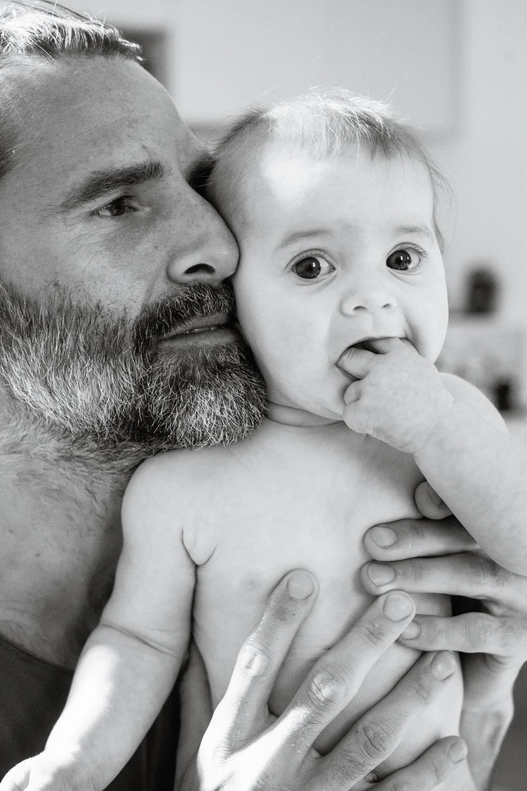 An older man with a beard holding a baby in his arms, with both of them close to each other. The baby has light hair, wide eyes, and is sucking on their fingers, looking at the camera. The image is in black and white.