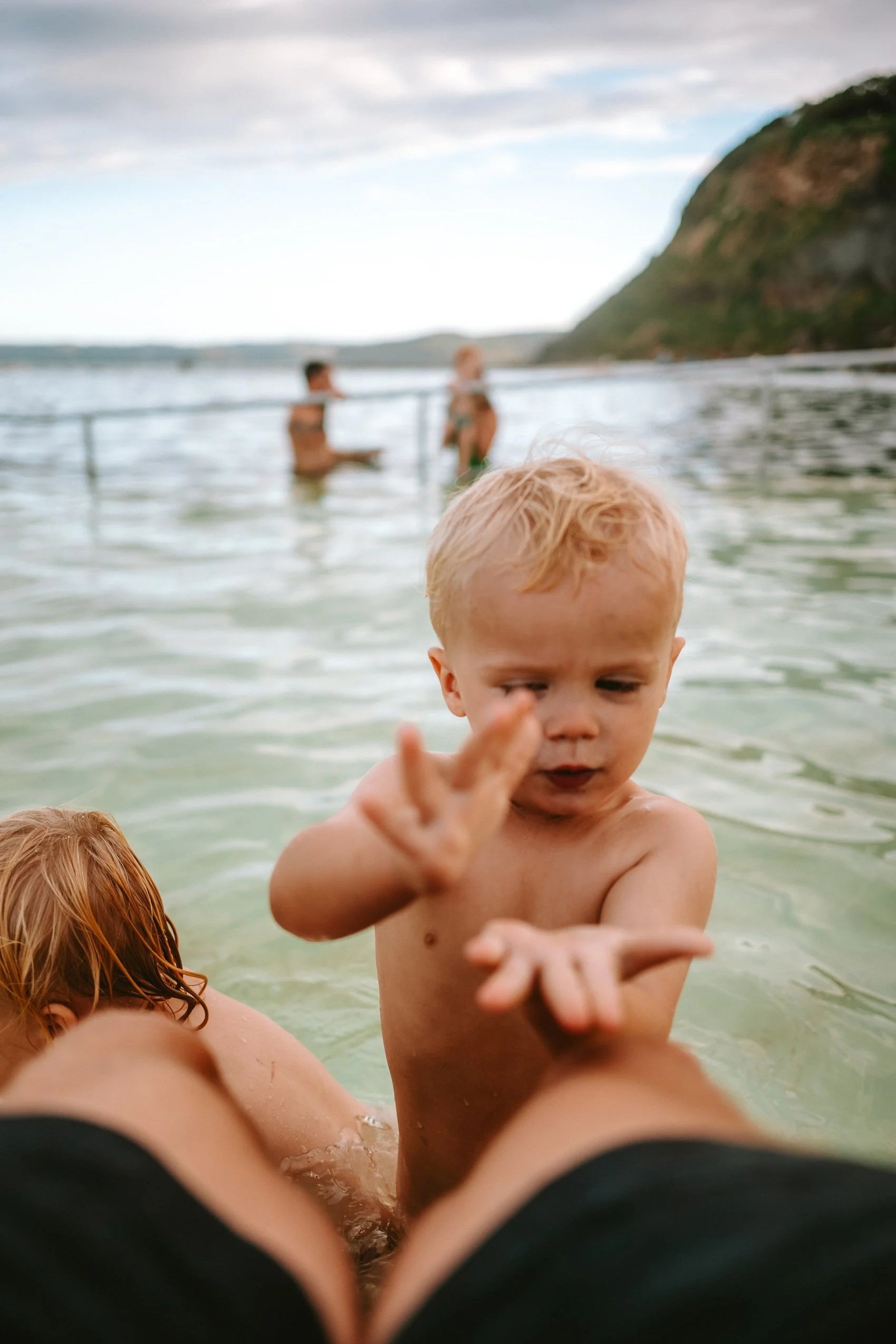 A young child with blonde hair reaching out with his hand while sitting in shallow water at the beach, with adults and hills in the background.