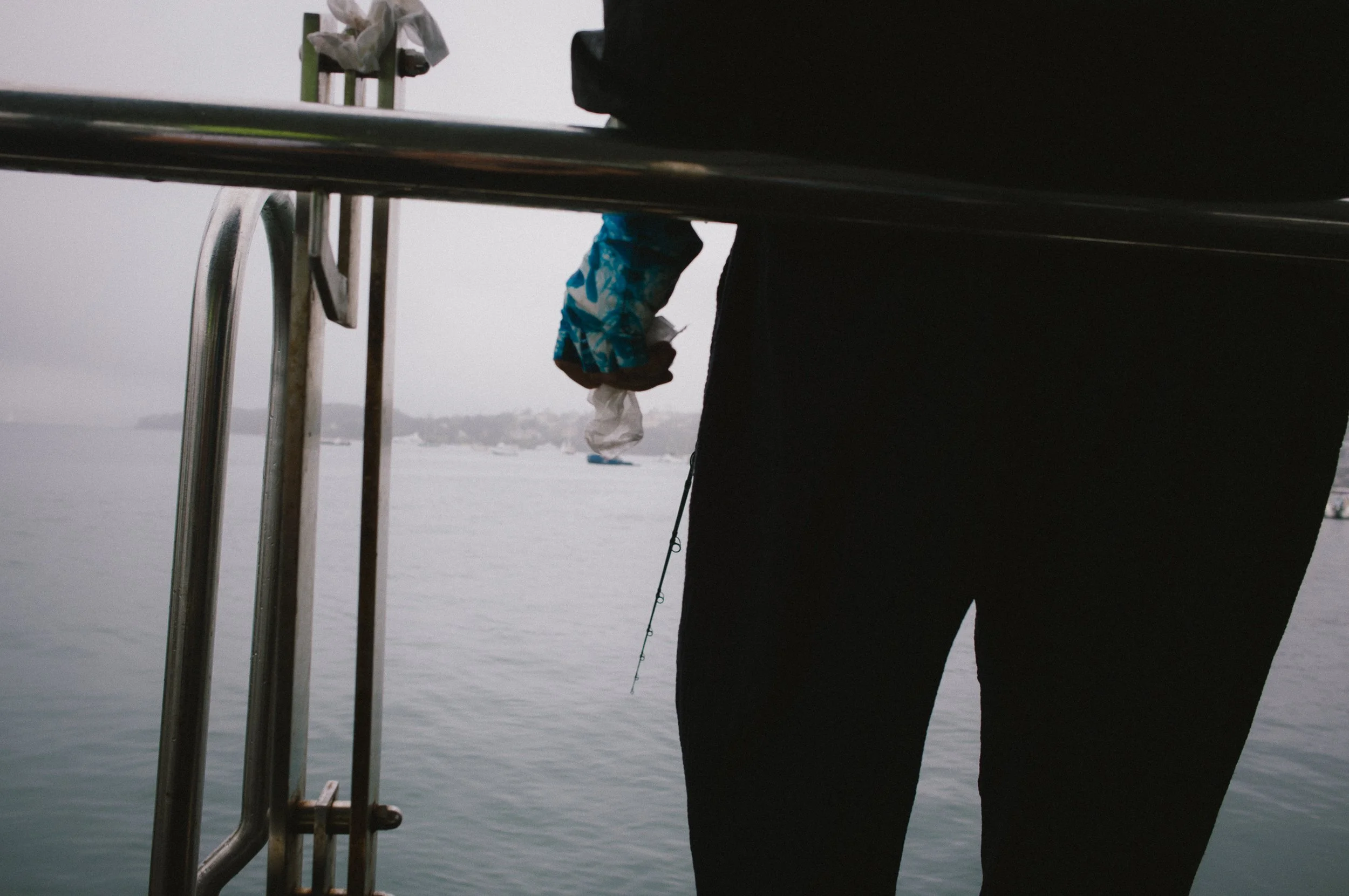 Person in black clothing standing near water, with a fishing rod and part of a metal structure in the foreground, overcast sky in the background.