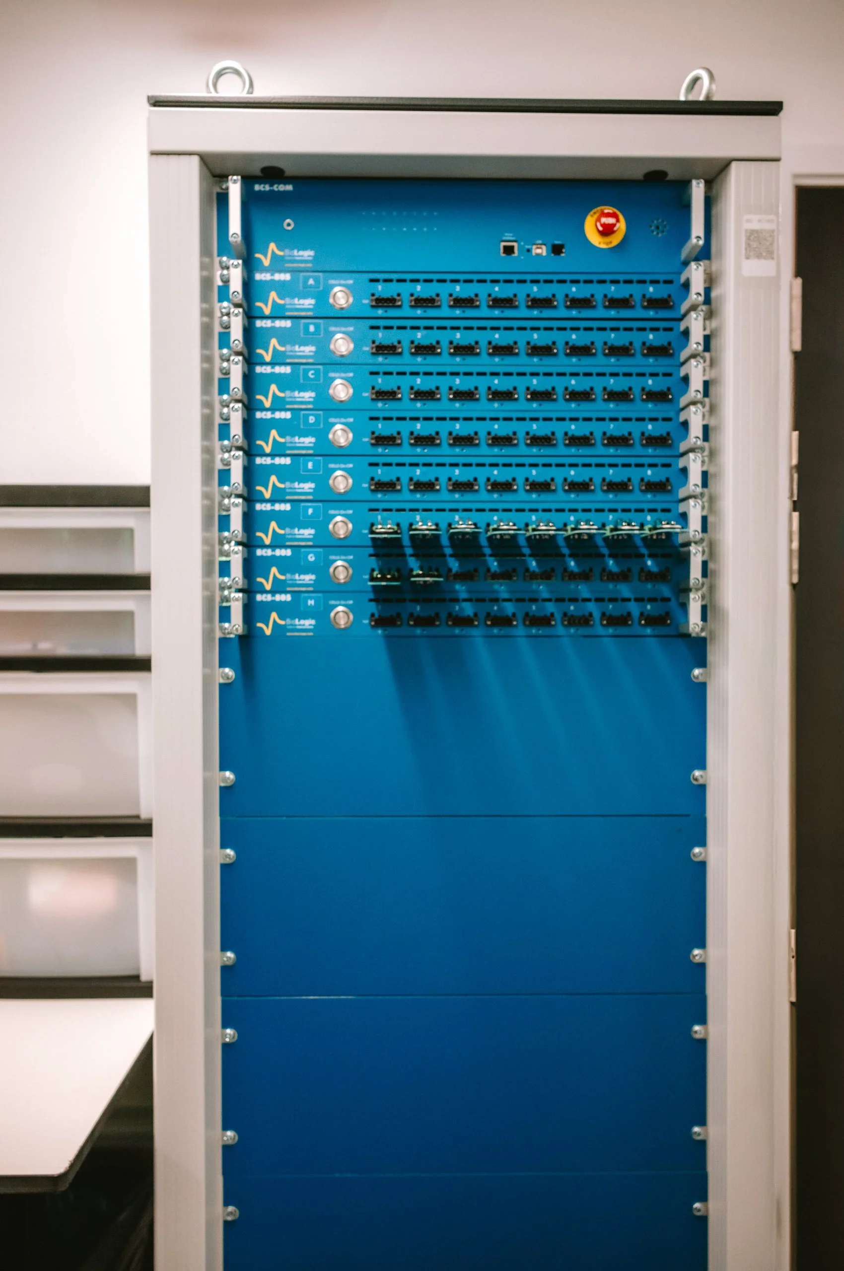 A medical equipment rack with multiple blue modules and black connectors, mounted in a white enclosure, with a red emergency stop button at the top right.