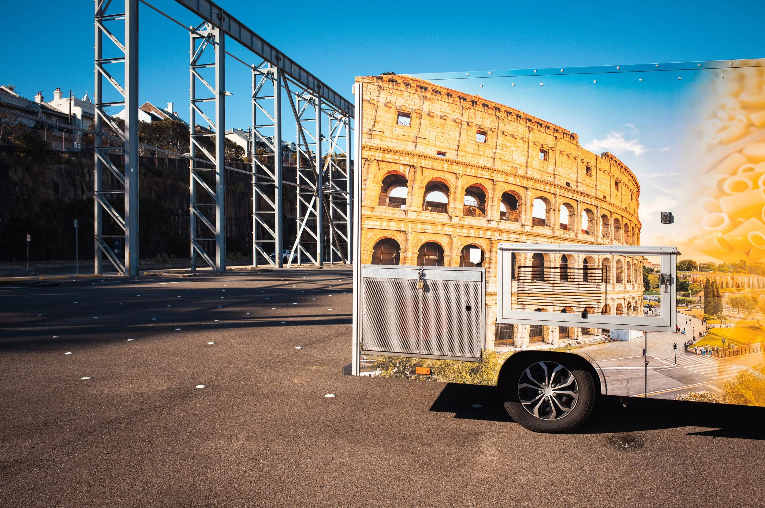 A food truck with an image of the Colosseum in Rome, Italy on its side, parked on an urban street with a construction site and a bridge in the background.