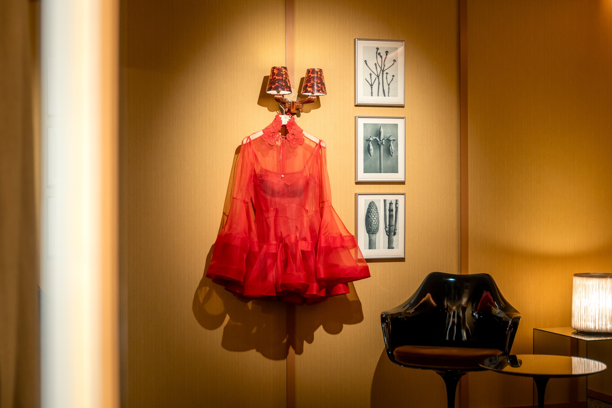 A red sheer dress hanging on a wall in a room with warm lighting, decorated with artwork, a black chair, and a side table with a lamp.