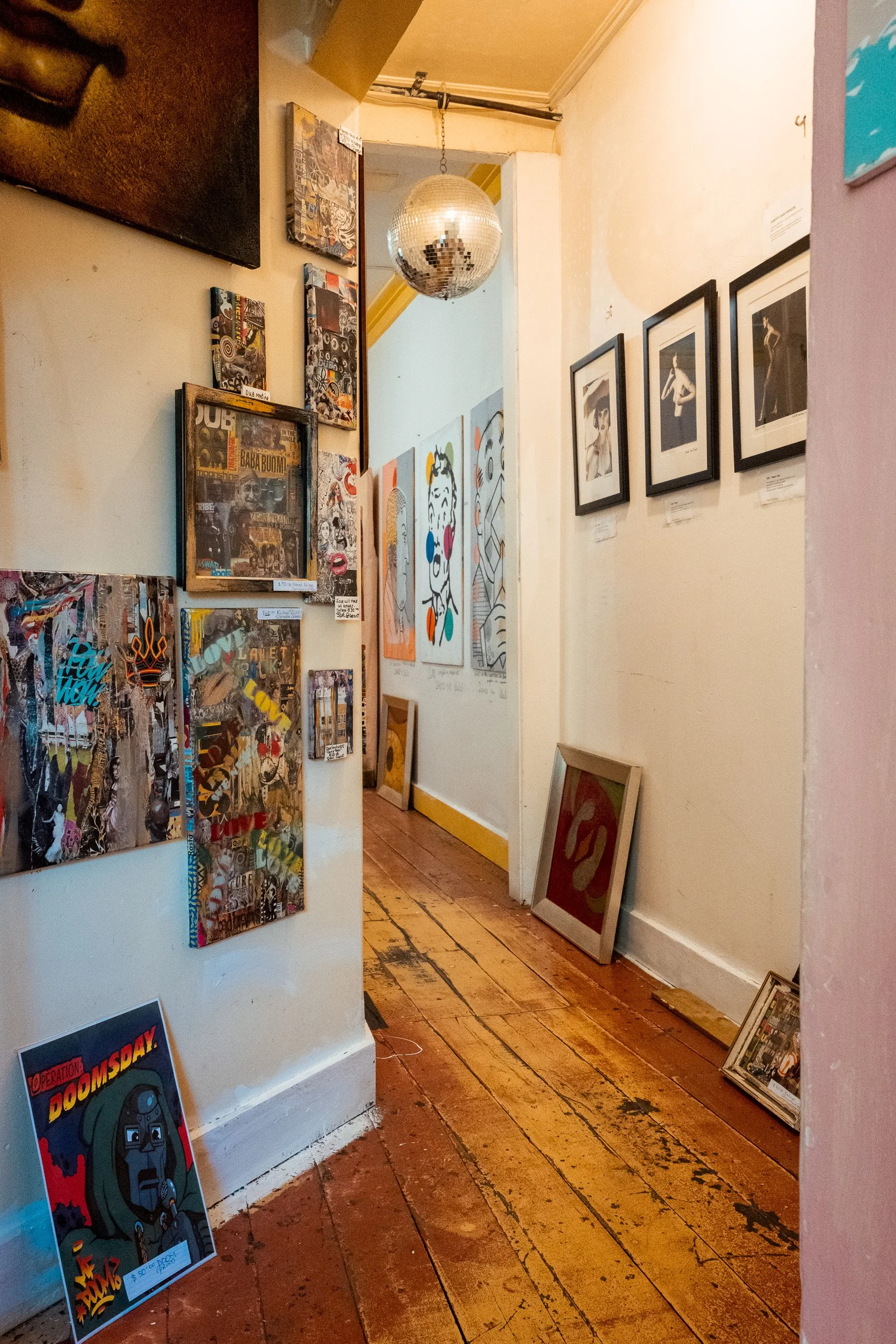 An art gallery hallway with paintings, photographs, and mixed media art displayed on the walls and leaning on the floor. A disco ball hangs from the ceiling, and the wooden floor shows signs of wear.