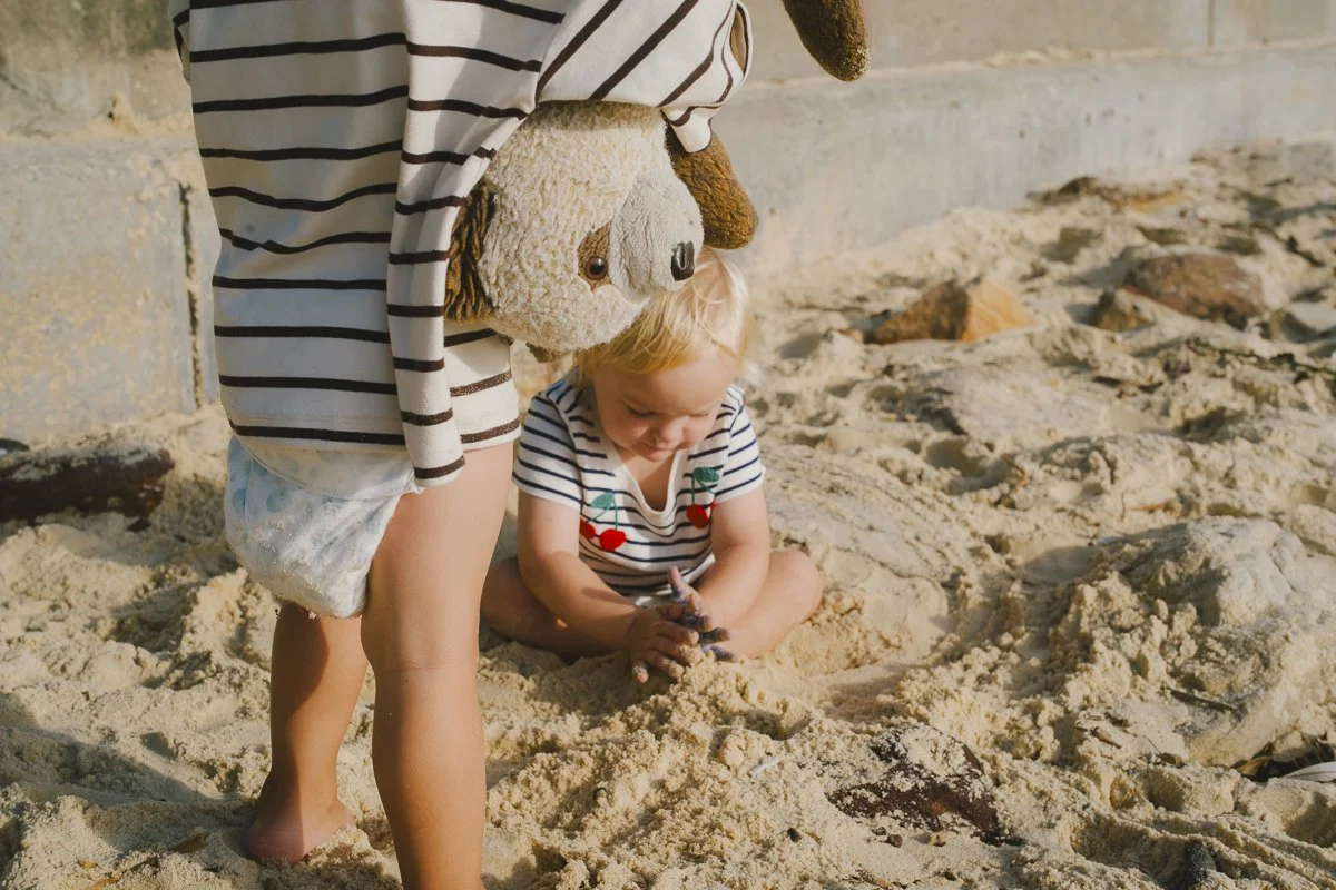 A young child playing in the sand with an adult, both wearing striped shirts. The adult is holding a teddy bear with a hat, and the child is digging in the sand.
