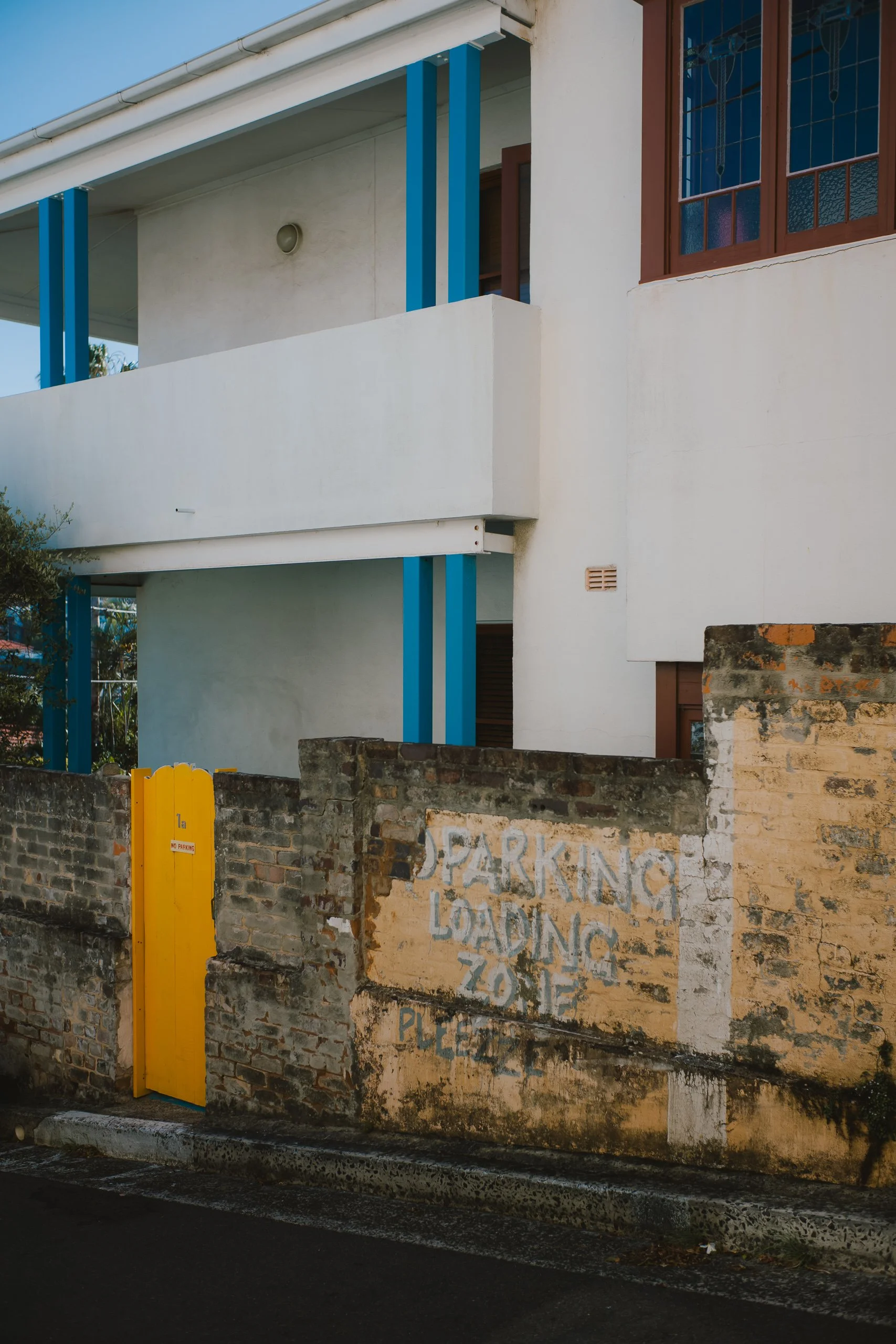 A white multi-story building with blue vertical supports and a brown window, behind a weathered brick wall with a yellow gate. The wall has faded graffiti that reads 'Parking Loading Zone Please'.