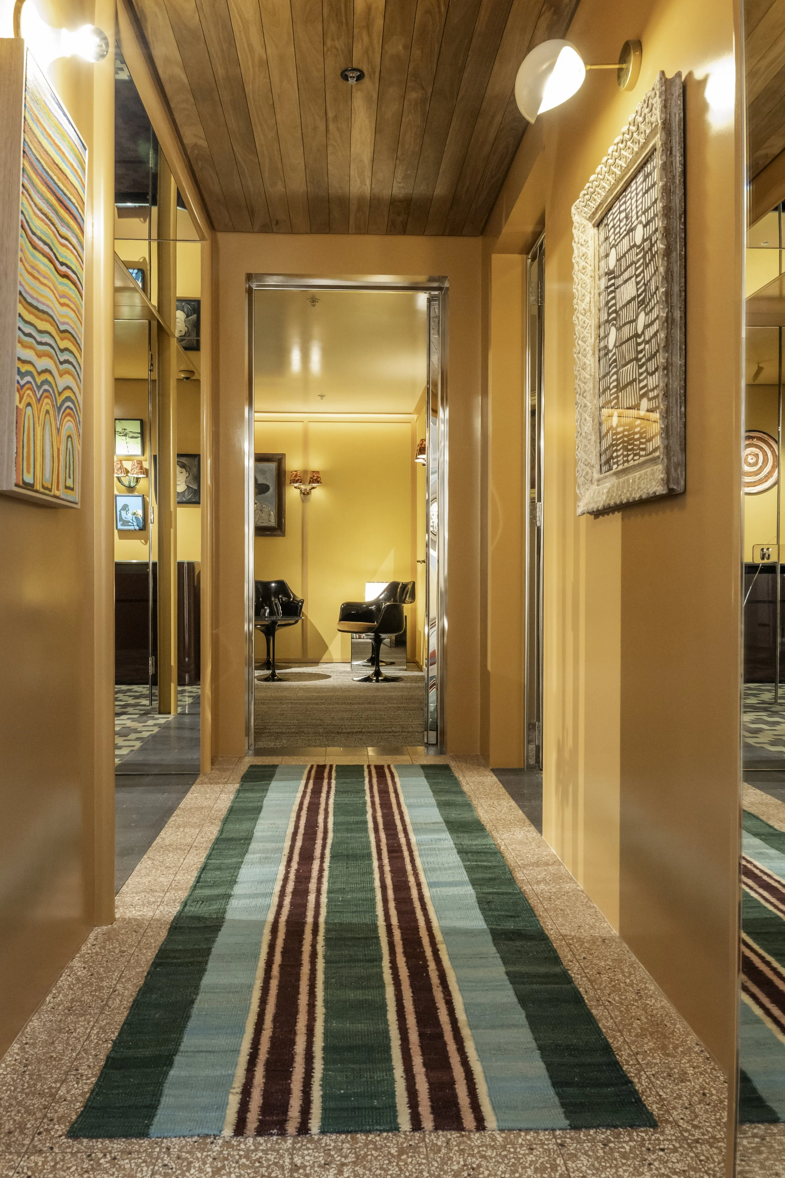 A hallway with orange from walls and ceiling, decorated with paintings and mirrors. The floor is covered with a colorful striped runner rug. At the end of the hallway, there is a seating area with two black chairs and a small table.