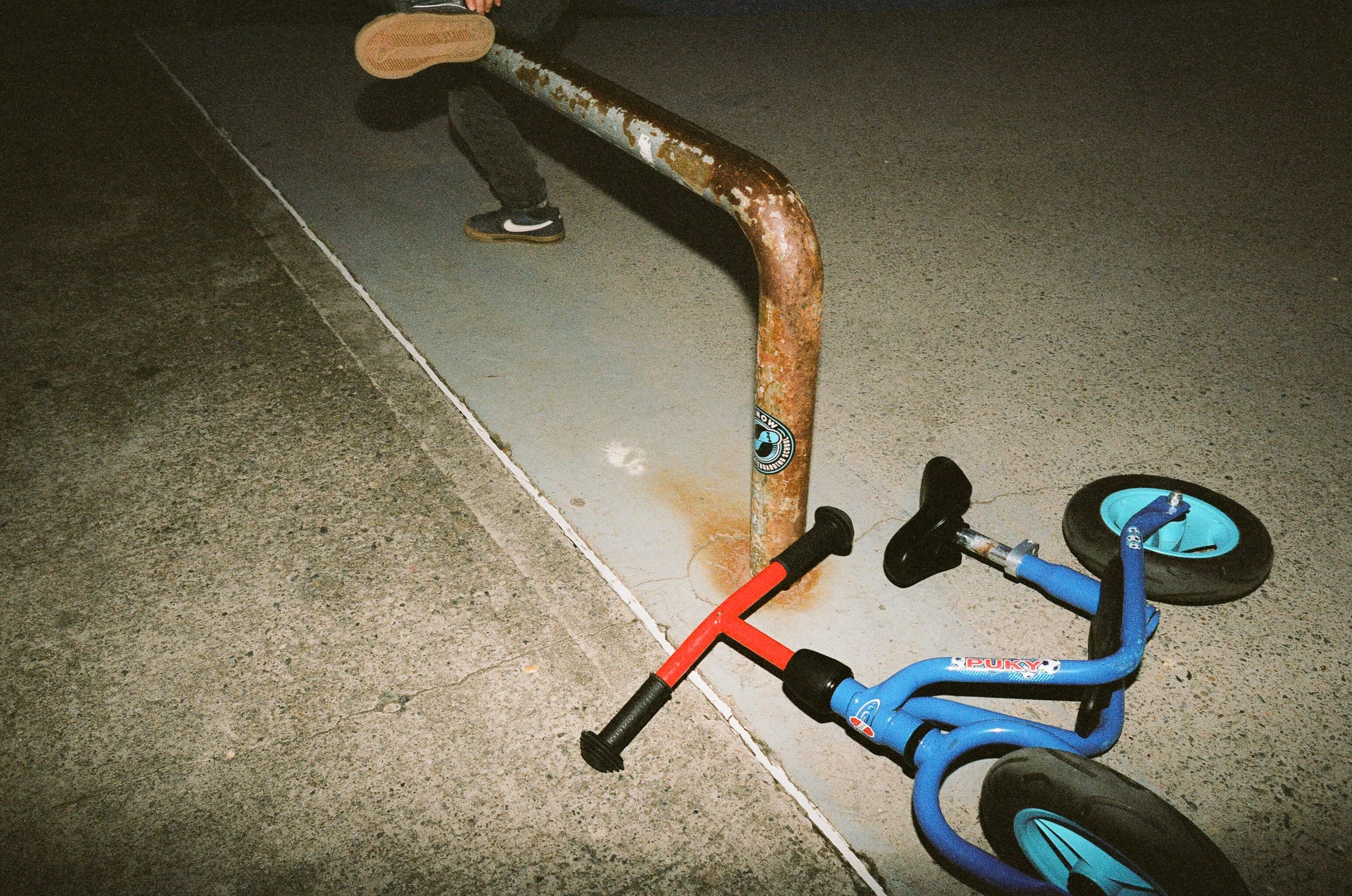 A rusty metal railing mounted on the sidewalk next to a child's blue and black balance bike with training wheels lying on the ground nearby.