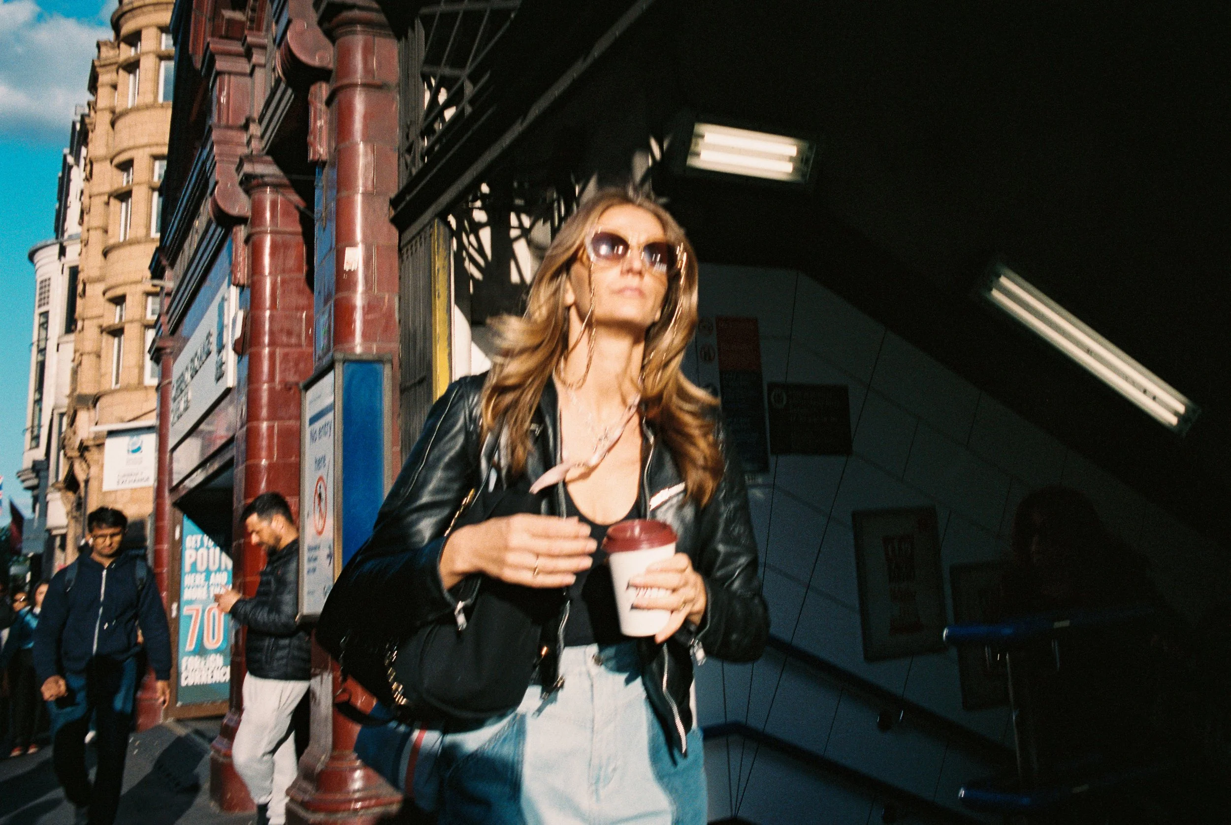 A woman with blonde hair, wearing sunglasses and a black leather jacket, holding a coffee cup, walking outside on a city street.