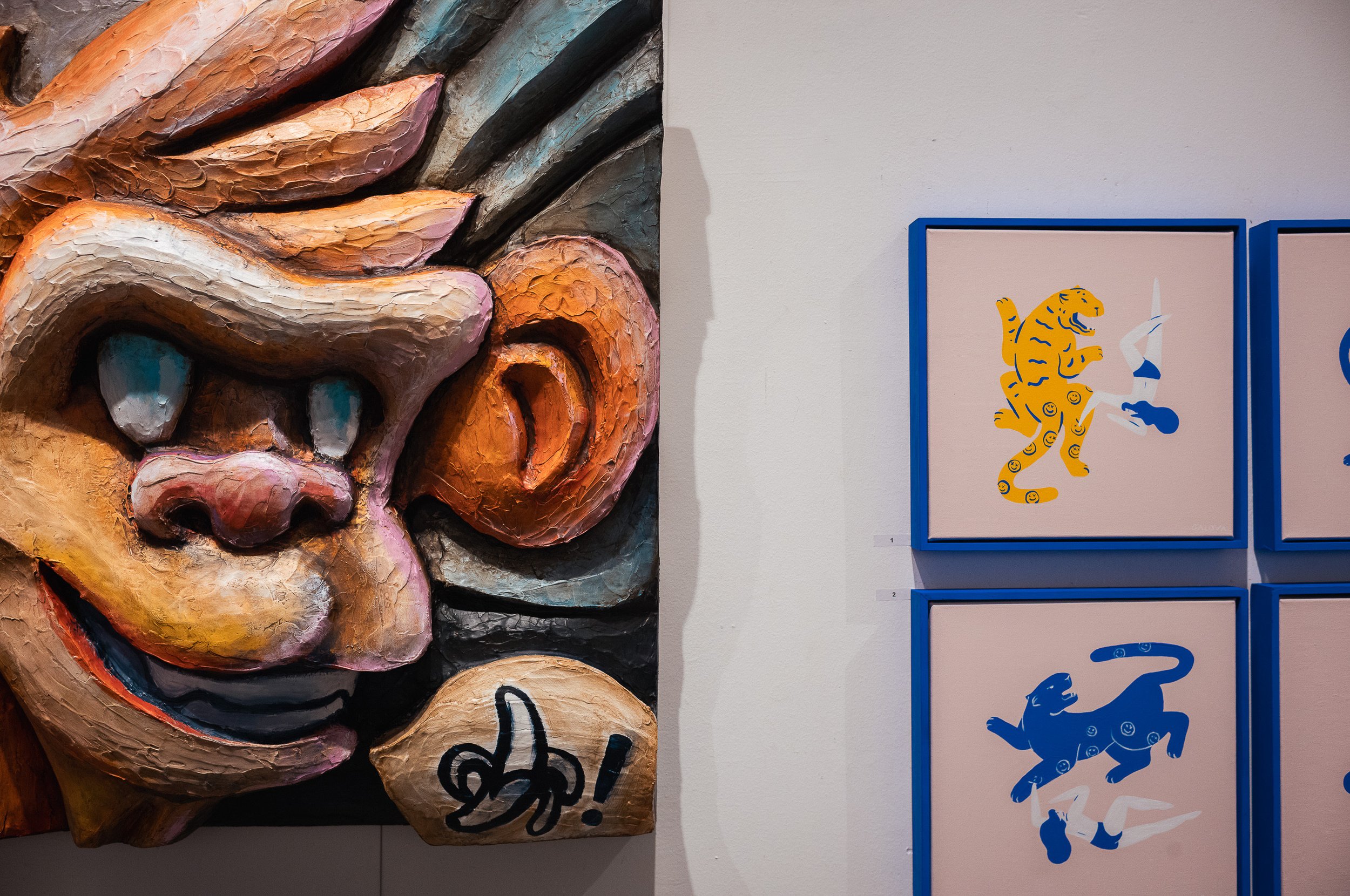 A large, colorful sculpture of a stylized lion's face with exaggerated features and textured surface, positioned next to framed artwork of stylized tiger and dog illustrations on a gallery wall.