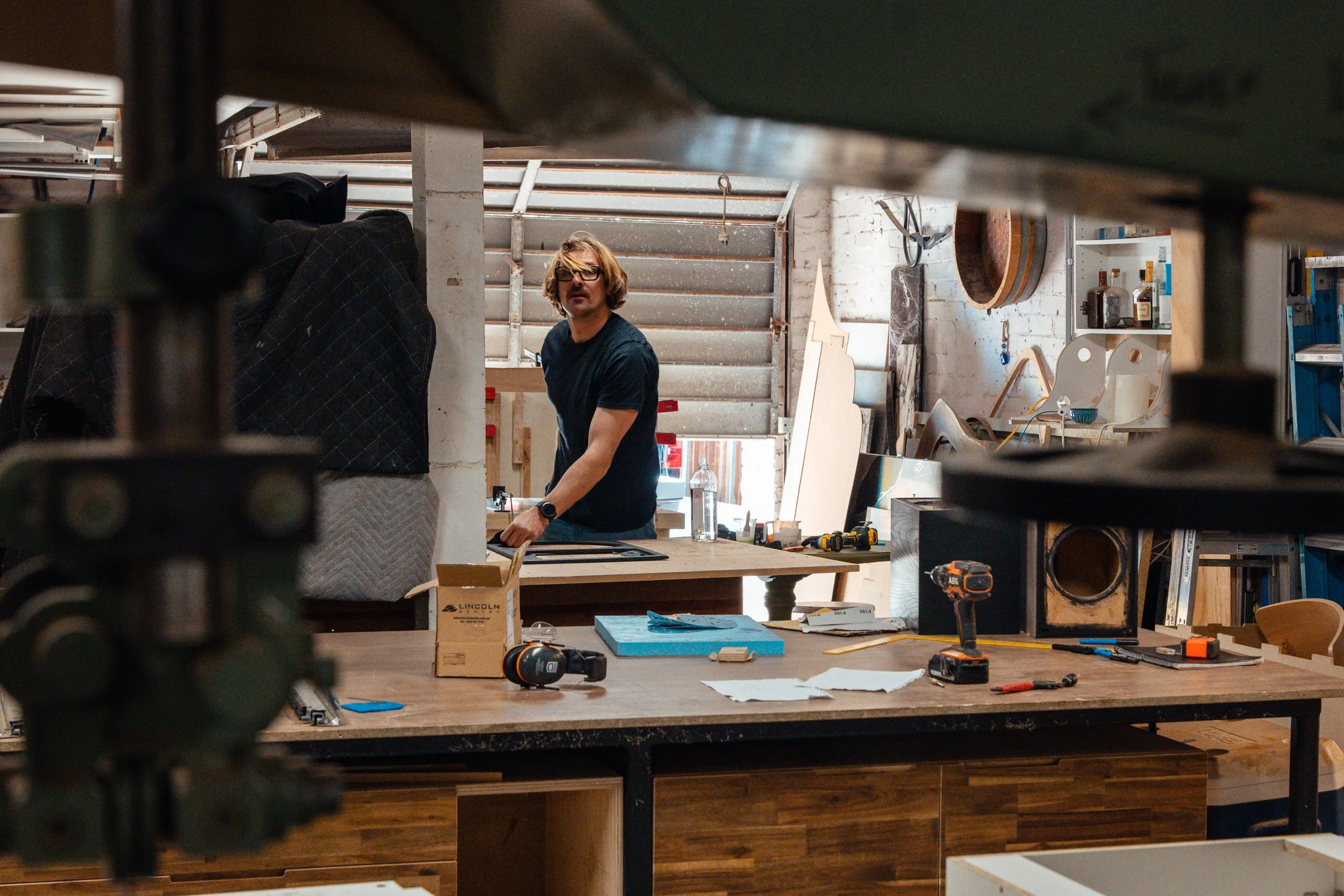 A man with glasses and a black t-shirt in a woodworking workshop, surrounded by tools and furniture in progress.