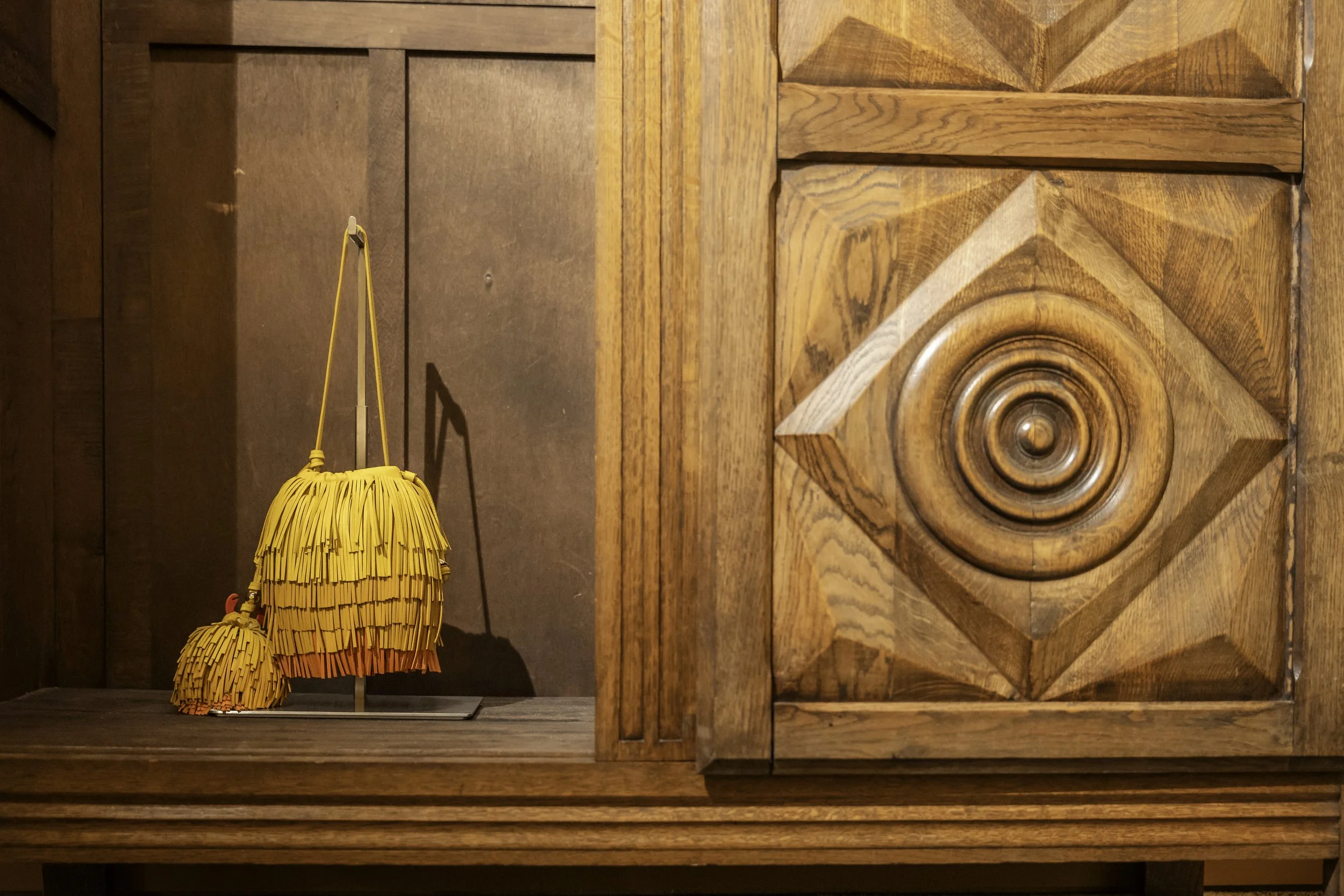 Two yellow handbags with fringe design, one larger and one smaller, are displayed on a wooden surface against a wooden wall with intricate patterns.
