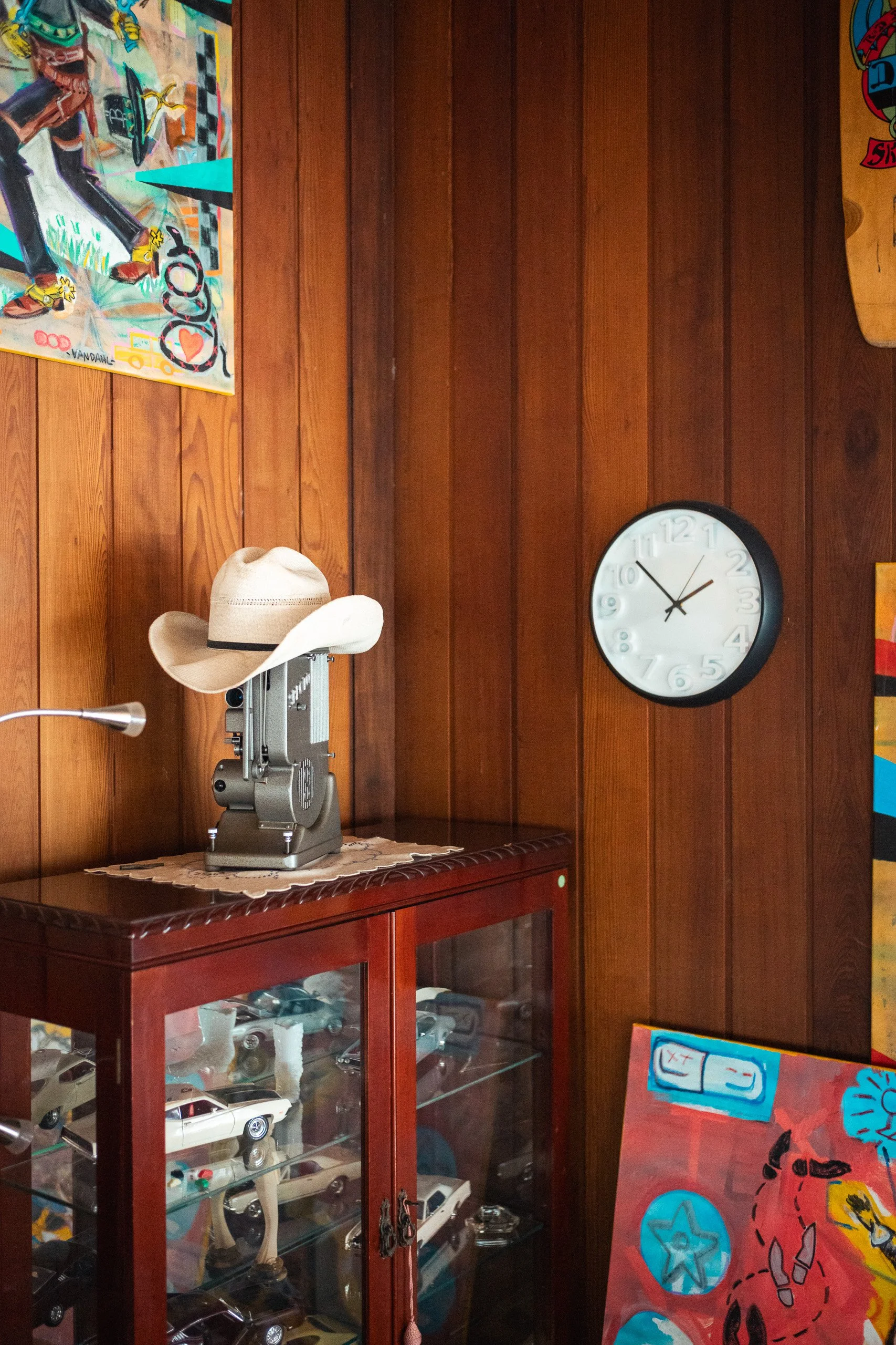 Wood-paneled room with a round clock on the wall showing 1:54, a display case filled with toy cars, a beige cowboy hat on a vintage film projector, colorful paintings on the walls, and a vintage desk lamp.