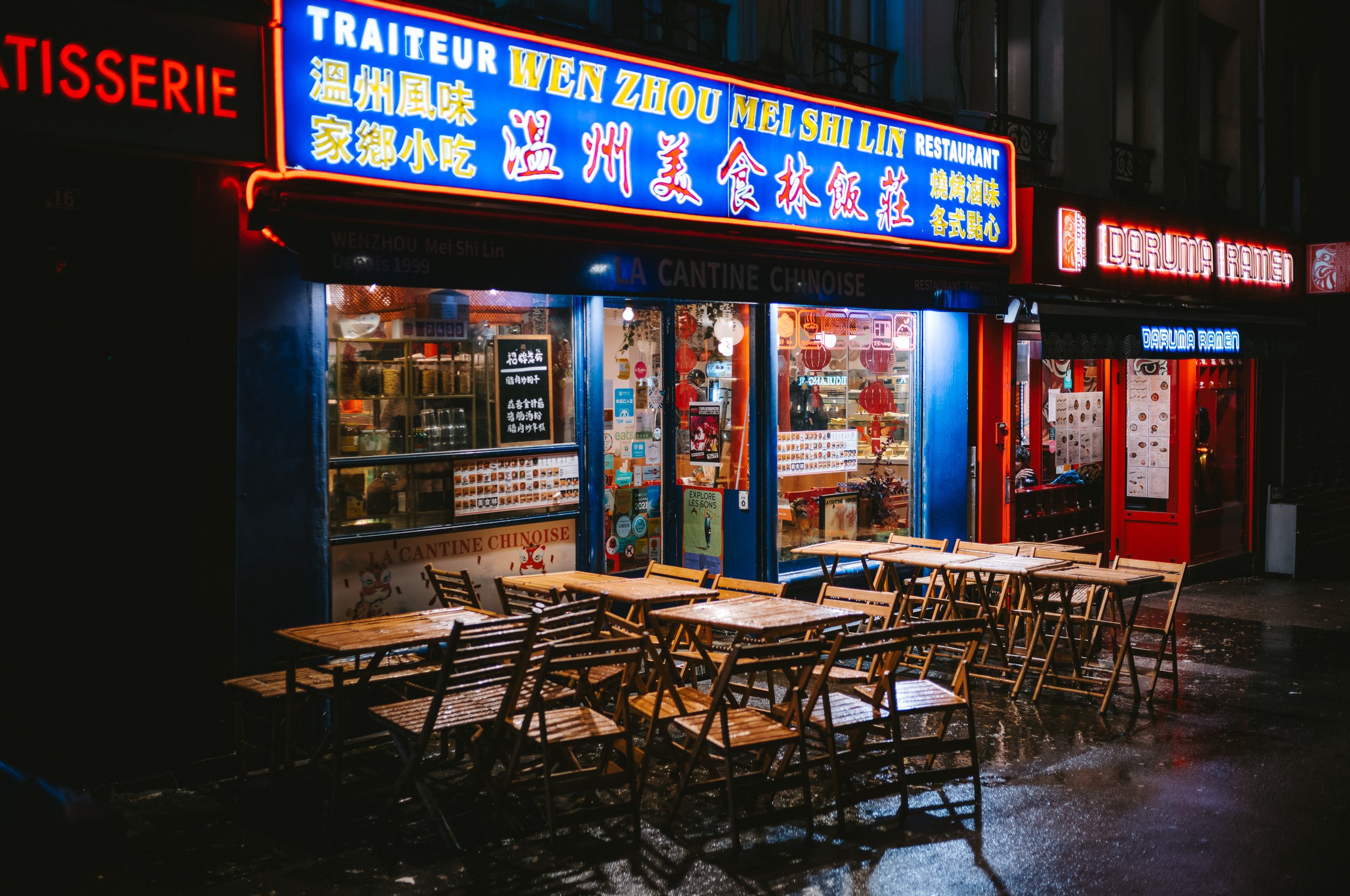 Nighttime street scene with a Chinese restaurant illuminated by neon signs, empty outdoor wooden tables and chairs, and a wet pavement reflecting lights.