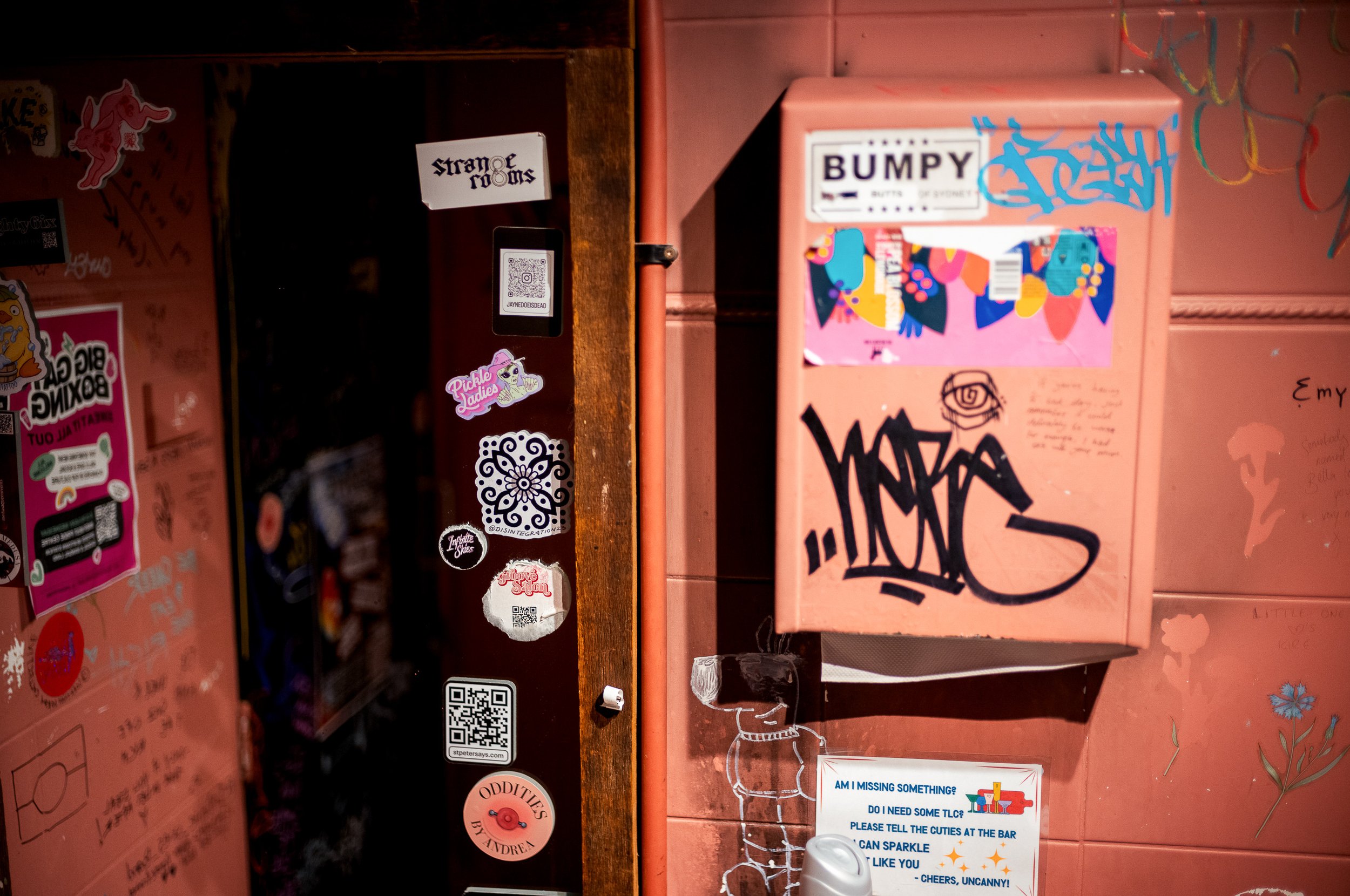 Pink wall with graffiti and various stickers, including a black graffiti tag, a sign that says 'BUMPY,' and a sign with a colorful graphic that reads 'WATERS.' There are also some small drawings, including a dog and other doodles.