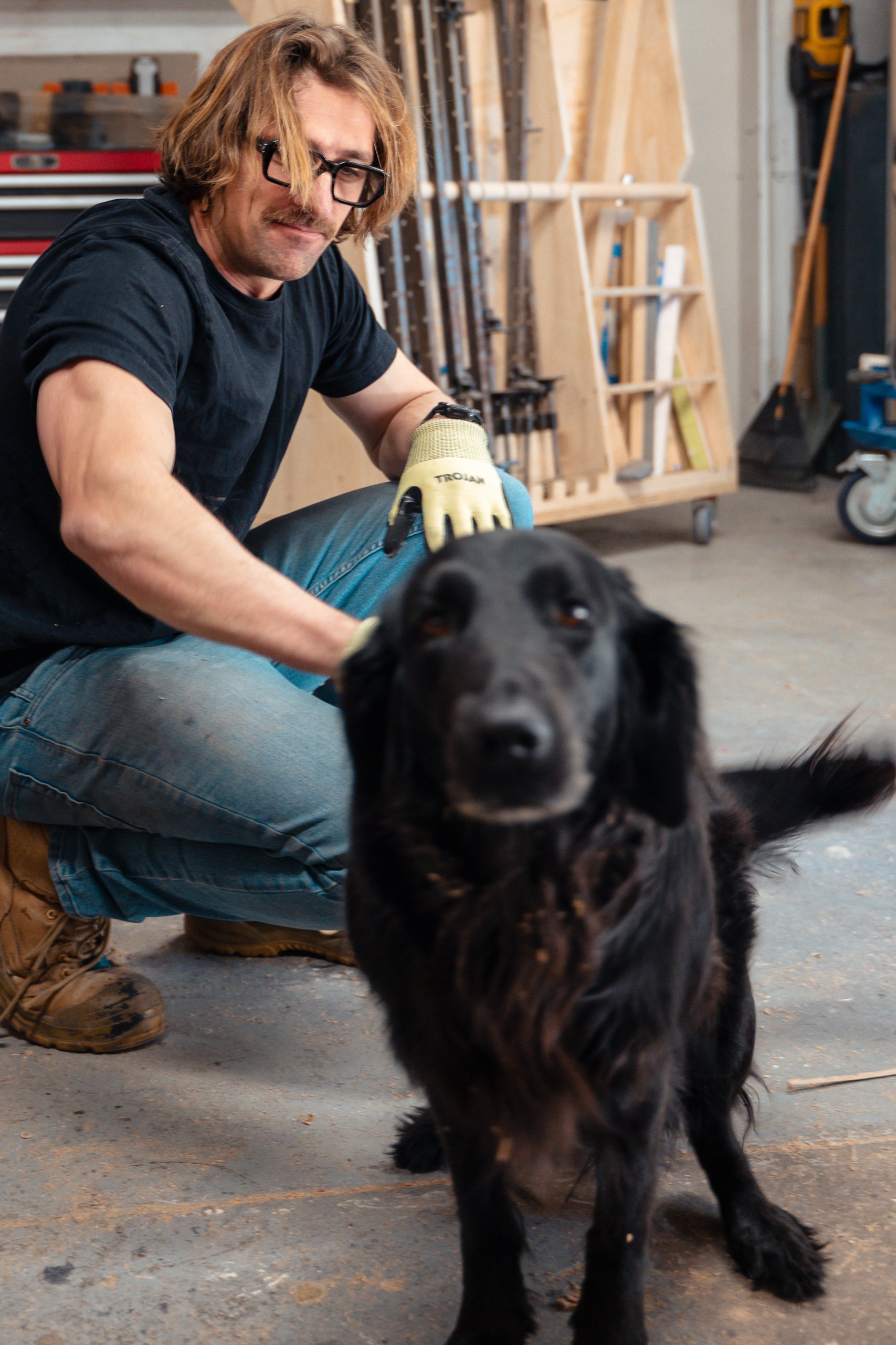 A man with glasses and long hair kneels in a workshop, tending to a black dog.