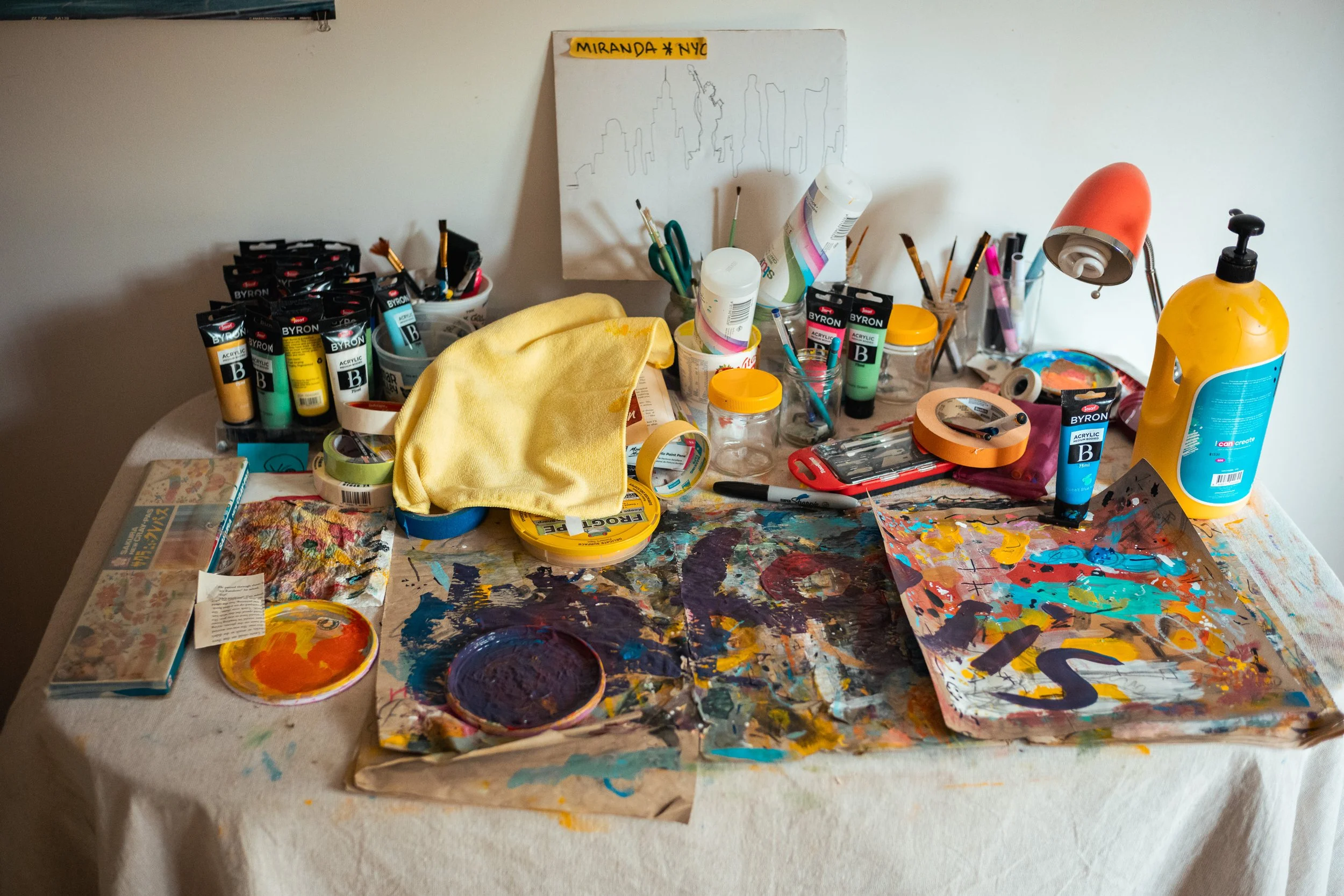 A messy art table with paints, brushes, tape, a yellow cloth, a small lamp, and a drawing of the New York City skyline on the wall.