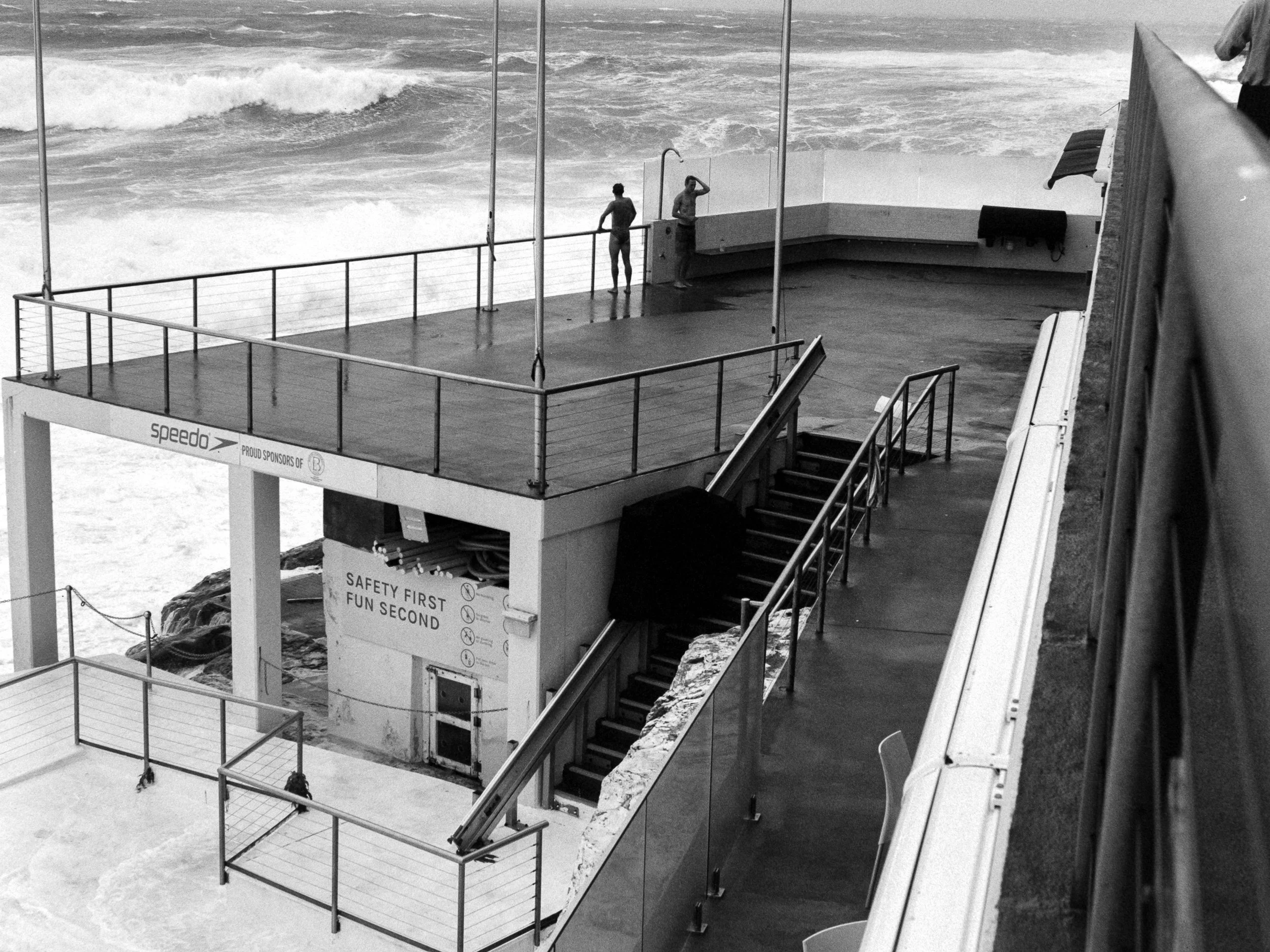 A black and white photo of a seaside deck with stairs and a railing, overlooking the ocean. Two people are standing on the deck near the water, one of whom appears to be adjusting their hair. There are signs with safety rules on the building below th