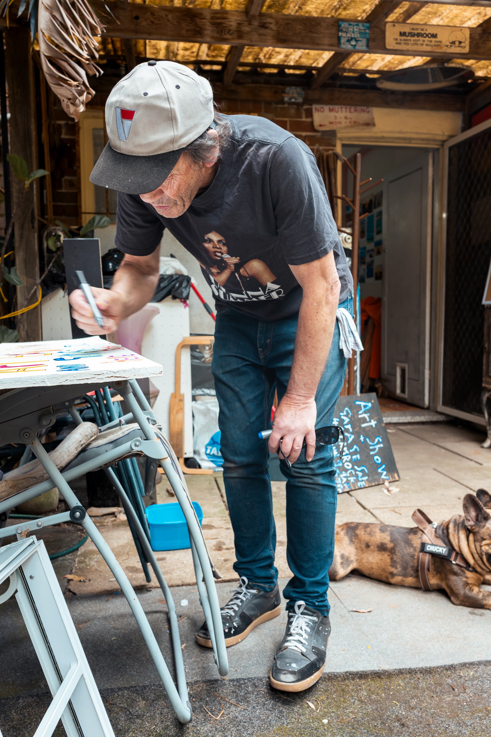 A man wearing a gray baseball cap, black t-shirt with a woman on it, and jeans is standing outdoors in a garage or shed, writing on a colorful sign or artwork on a table. A brown brindle dog with a collar is lying nearby on the ground.