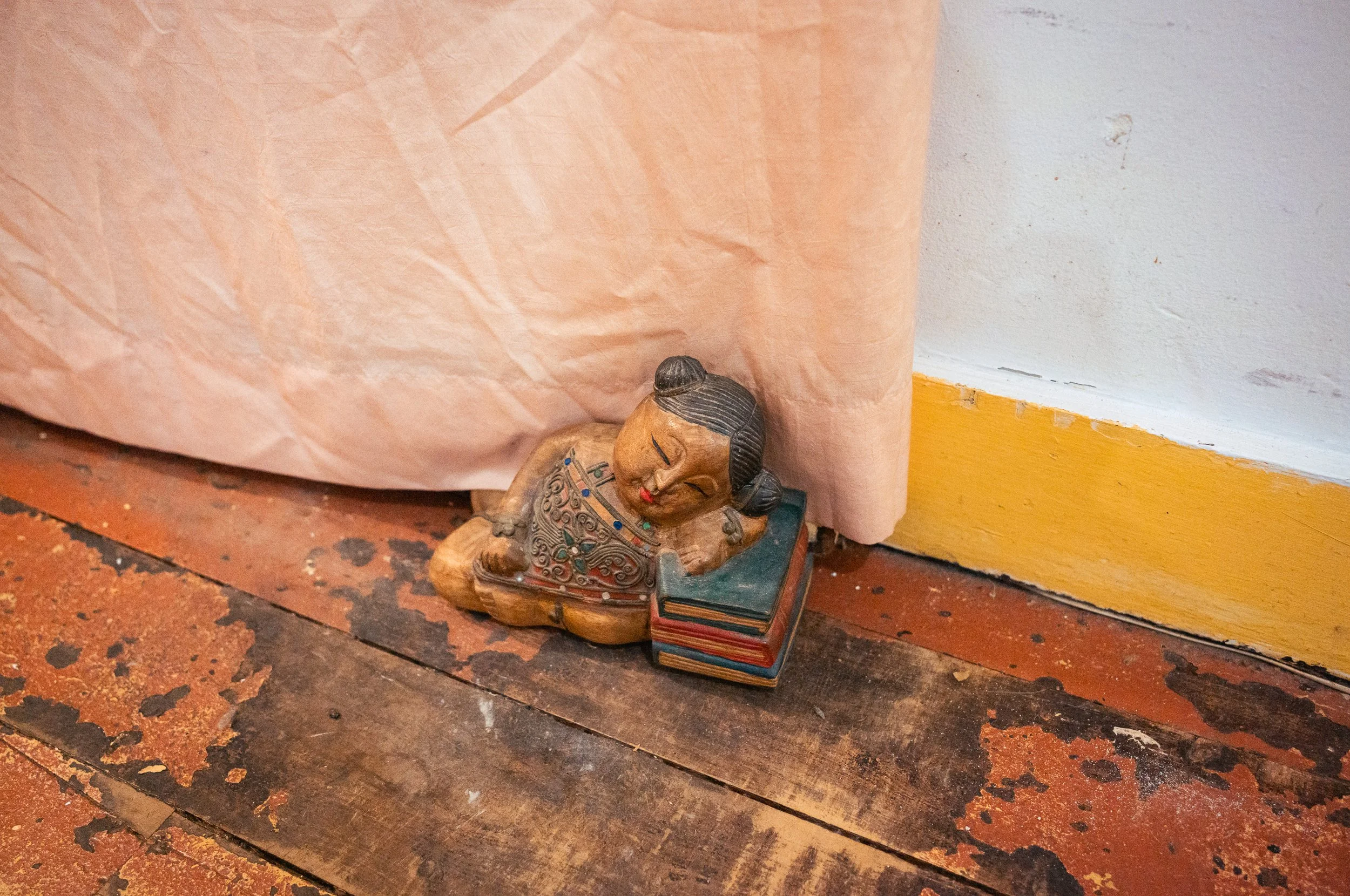 A small carved wooden figurine of a woman in traditional dress lying on the floor next to a pink curtain and a wooden baseboard, with a stack of small books underneath her.