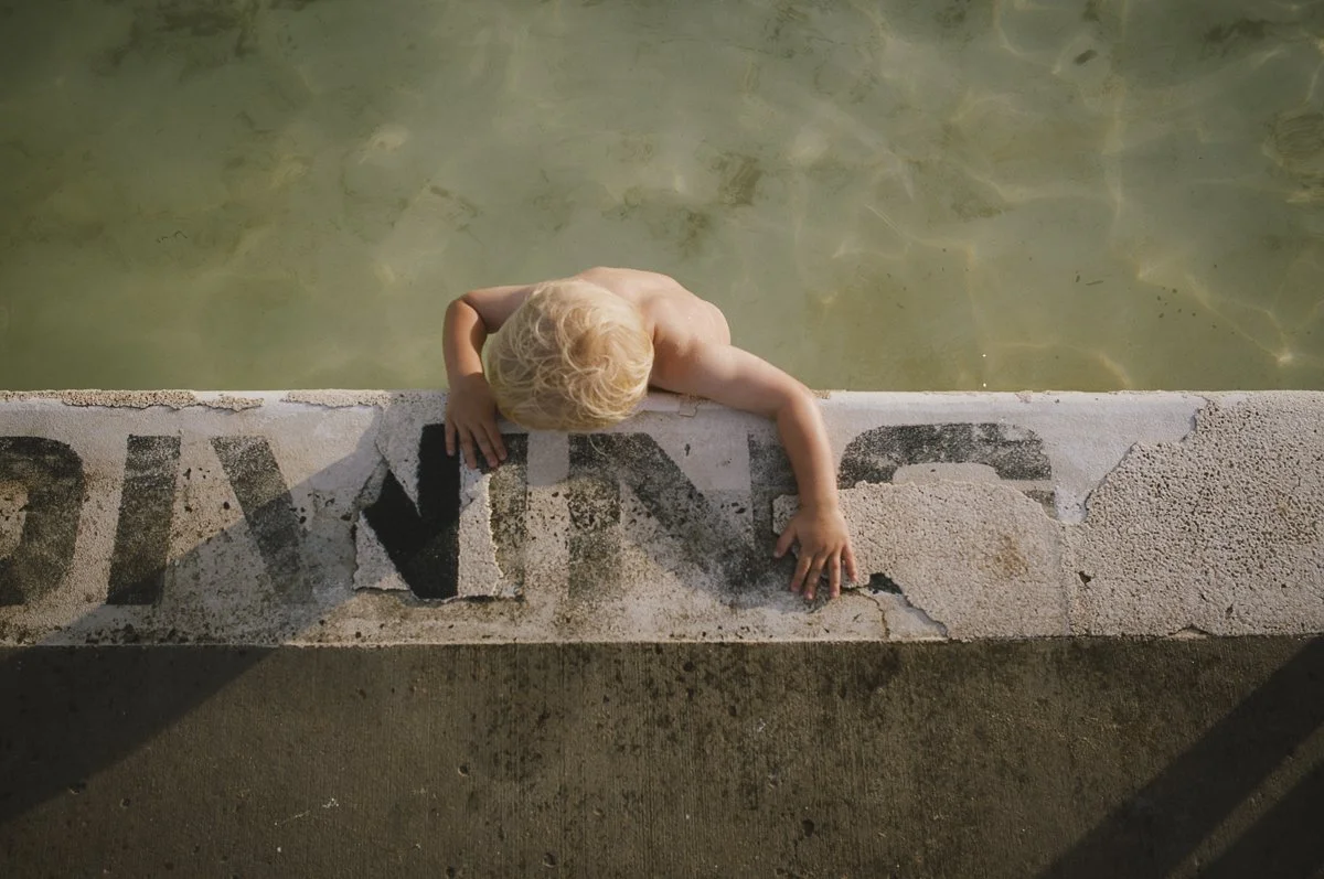 Young child with blond hair leaning over the edge of a concrete poolside, reaching into the water.